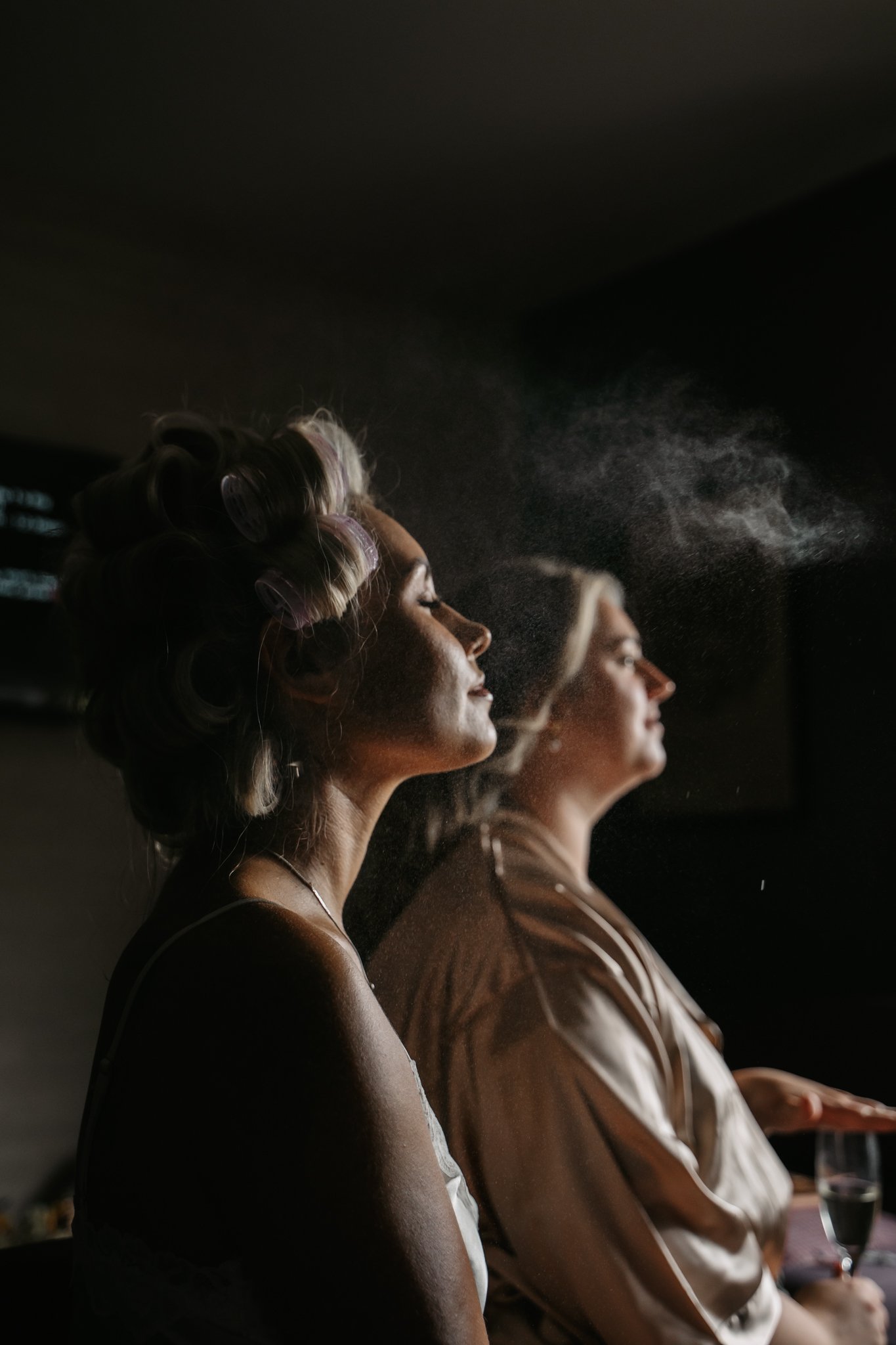 Two women sitting side by side with their eyes closed, one with hair rollers and the other with a headband, in a dimly lit room, with the first woman spraying perfume.