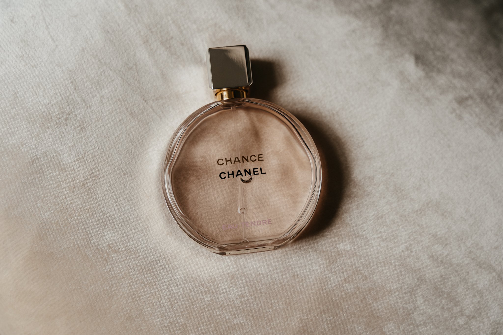 A round glass bottle of Chanel Chance Eau Tendre perfume with a silver cap resting on a silky beige fabric.