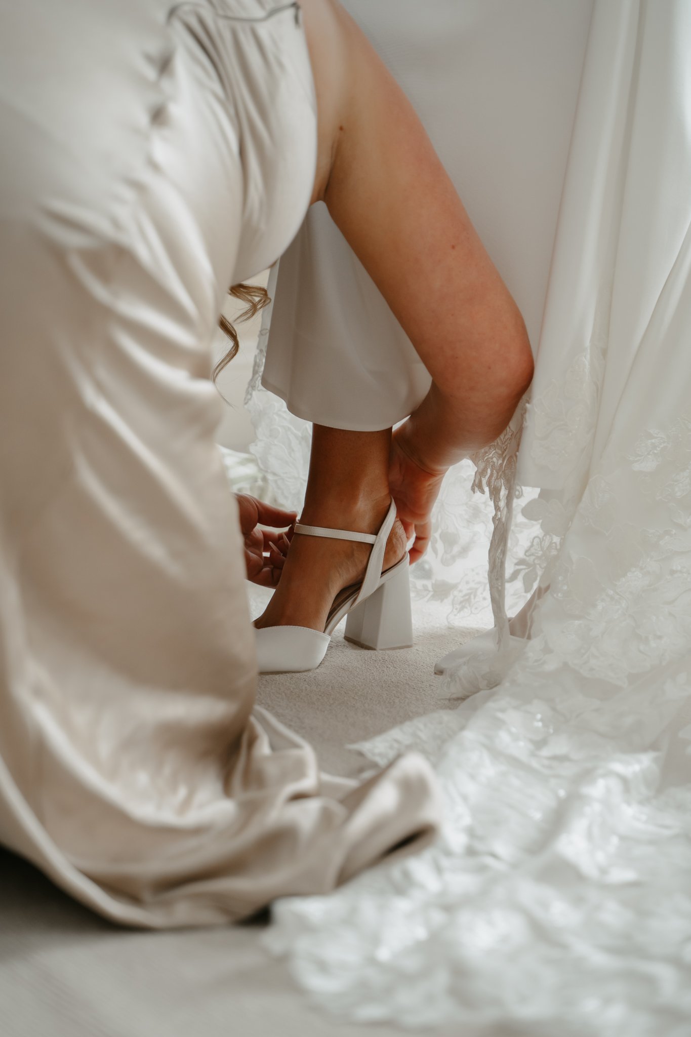 A woman in a cream-colored satin robe adjusts her white high-heeled shoe on her foot as she prepares for a wedding or formal event, with lace curtains and a patterned dress or gown visible.