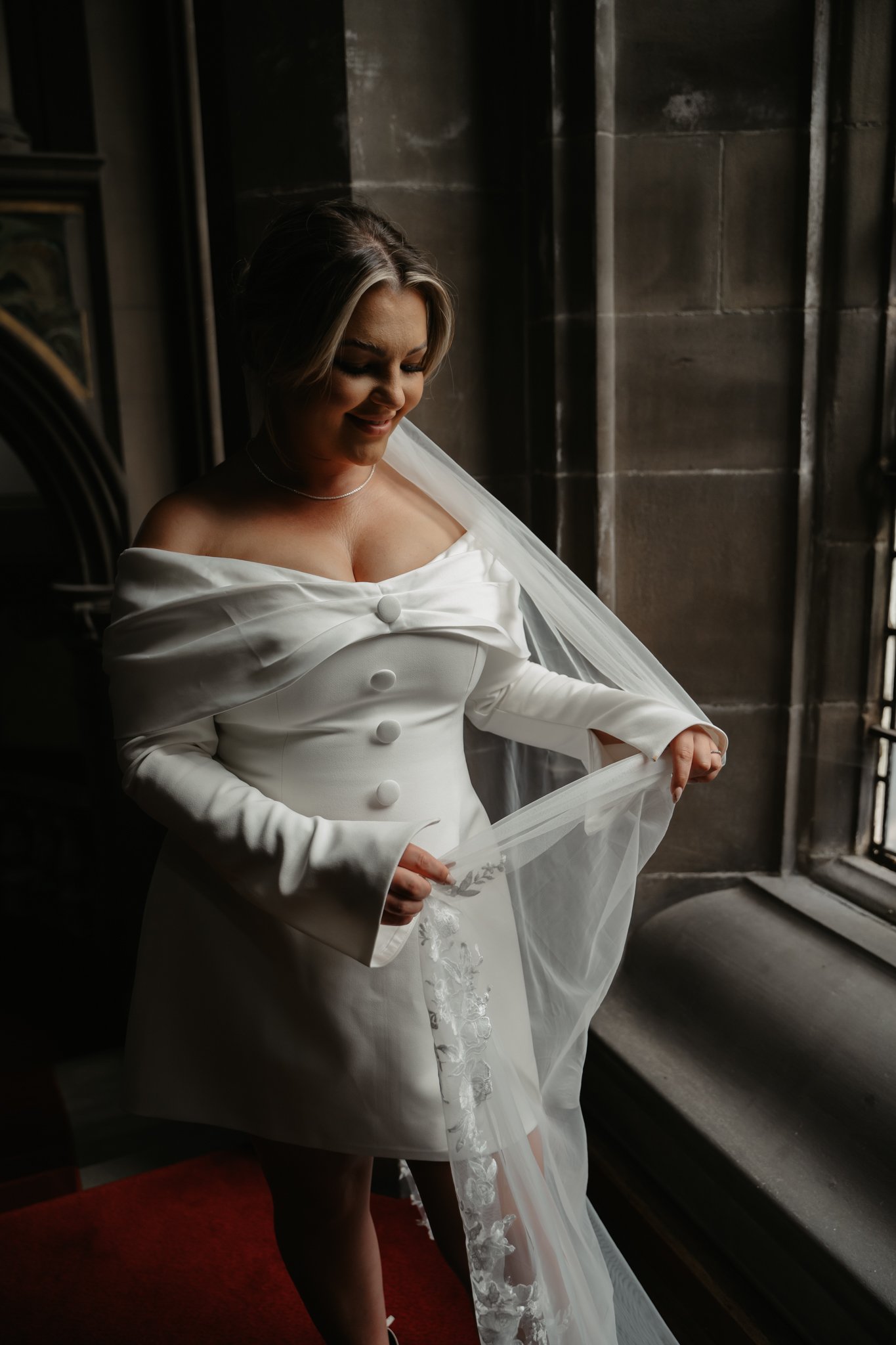 A bride in a white dress holding a veil near a window in a stone building.