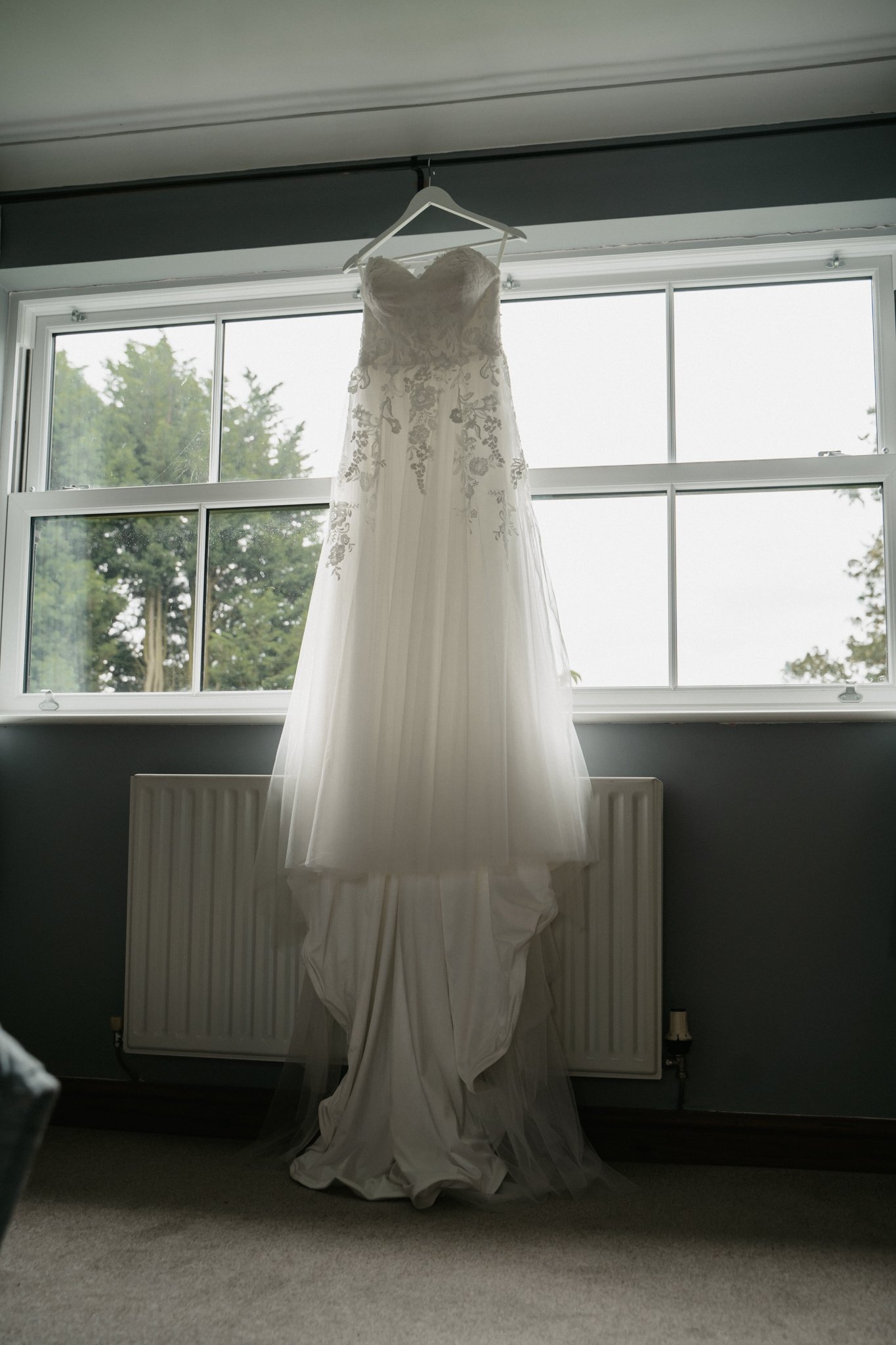 A wedding dress hanging from a white hanger in front of a window with trees outside.