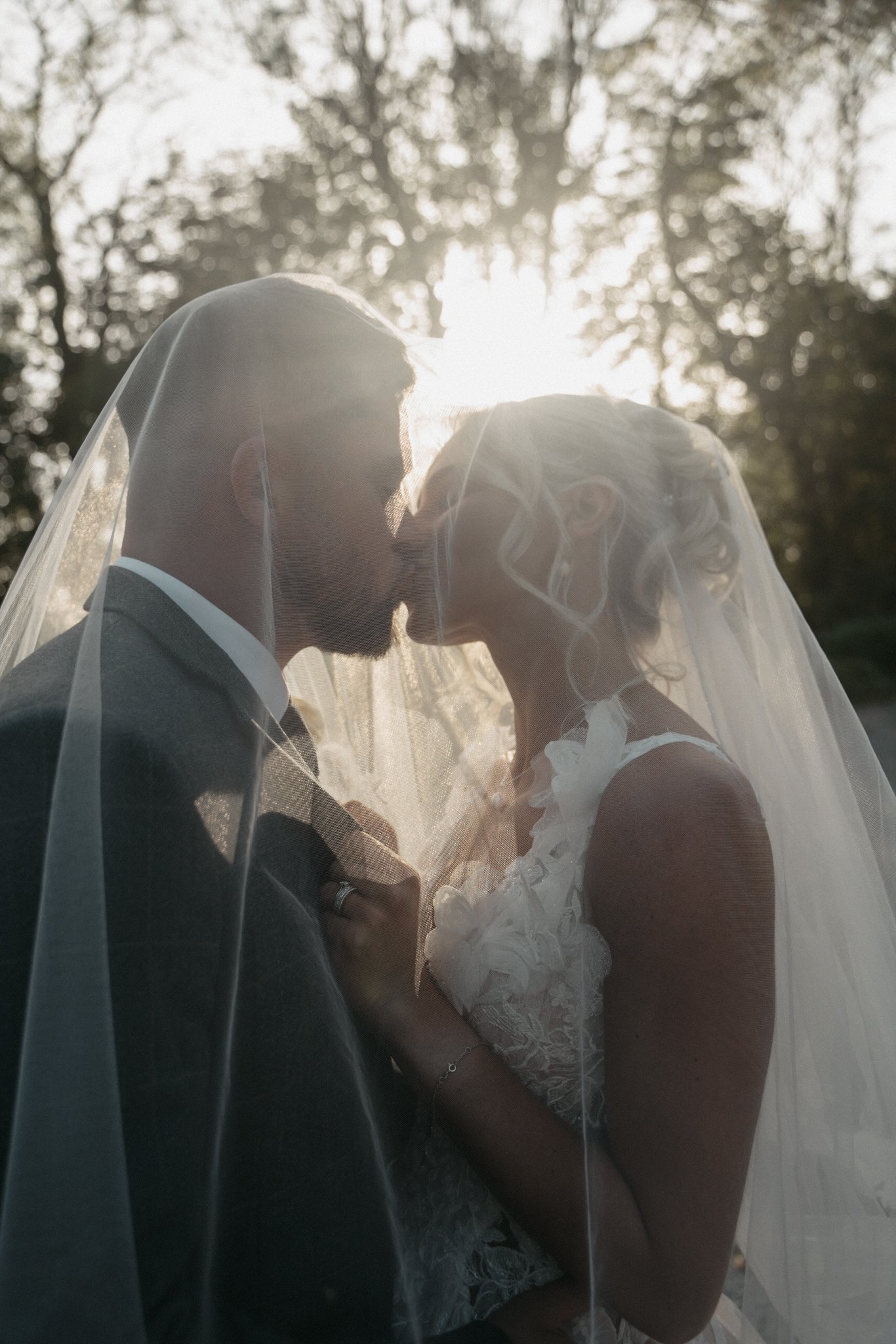 A bride and groom sharing a kiss under a veil outdoors with sunlight behind them.