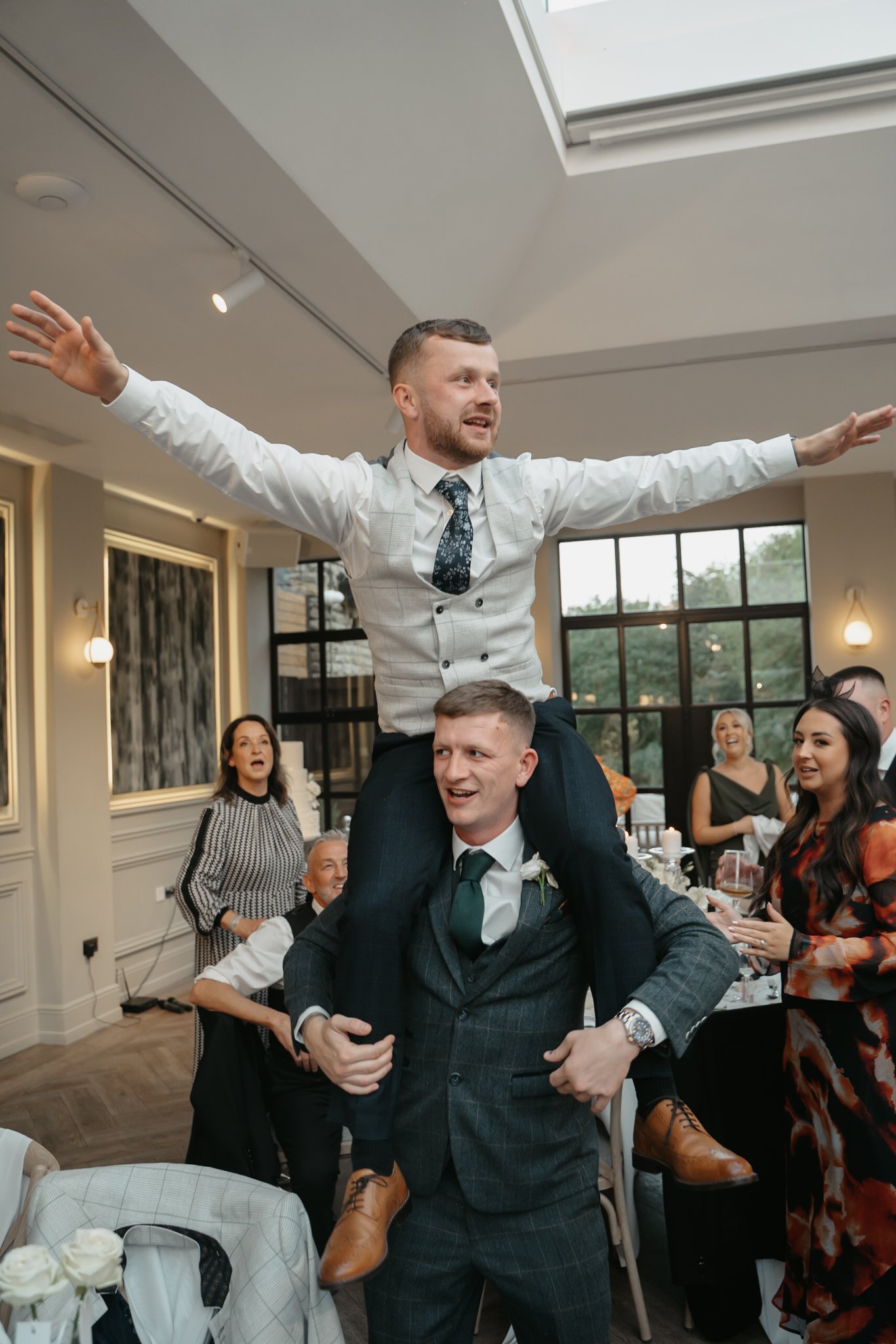 A man in a suit is giving another man a piggyback ride, who is standing with his arms outstretched, at a wedding reception. Several people are standing and sitting around, smiling and watching.