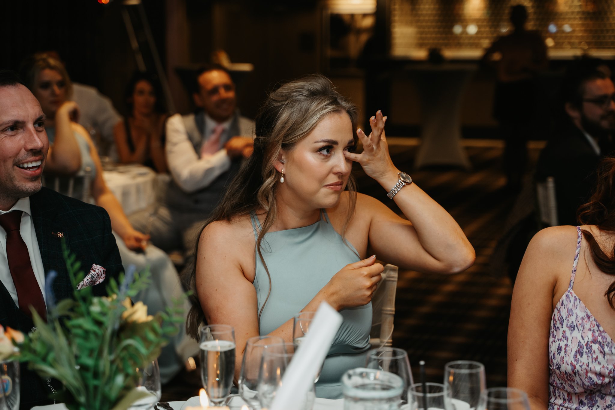 Woman wiping tears from her eyes at a formal event, surrounded by seated guests, some smiling and clapping.