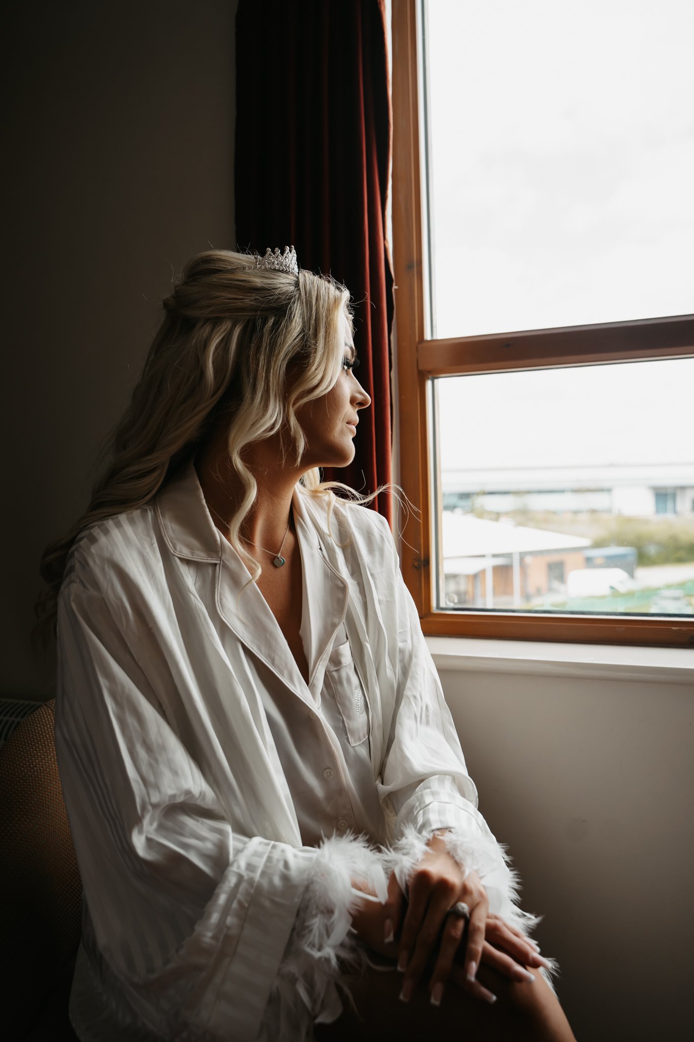 A woman with long blonde hair, wearing a small tiara and a white satin robe with feathered cuffs, sits by a window looking outside. She has a necklace with a blue pendant and a ring on her finger.