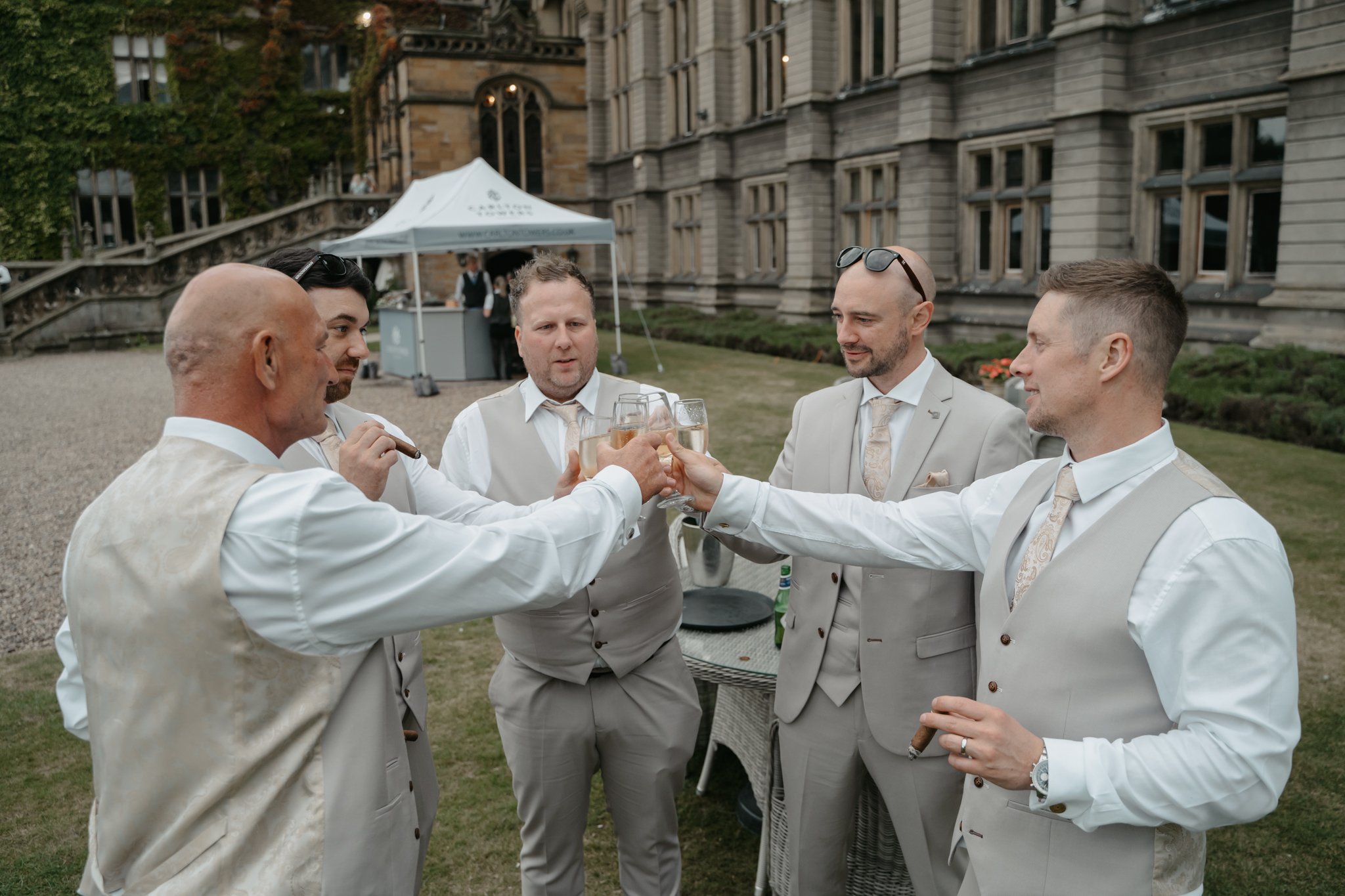 Five men in suits and vests celebrating with drinks outdoors in front of a historic building, with a table and tent in the background.