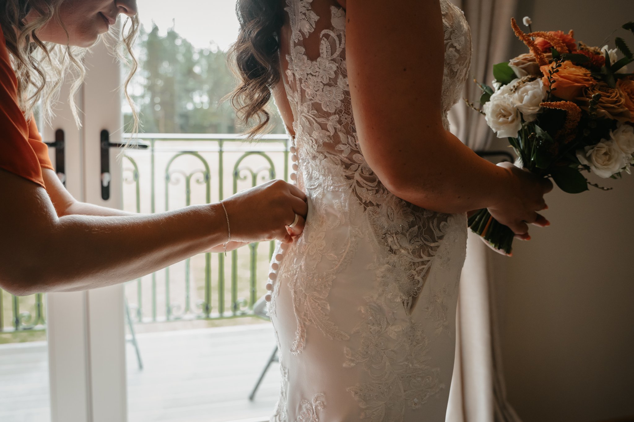 A bride is getting help fastening her wedding dress as she holds a bouquet of flowers, standing near a window with a balcony in the background.