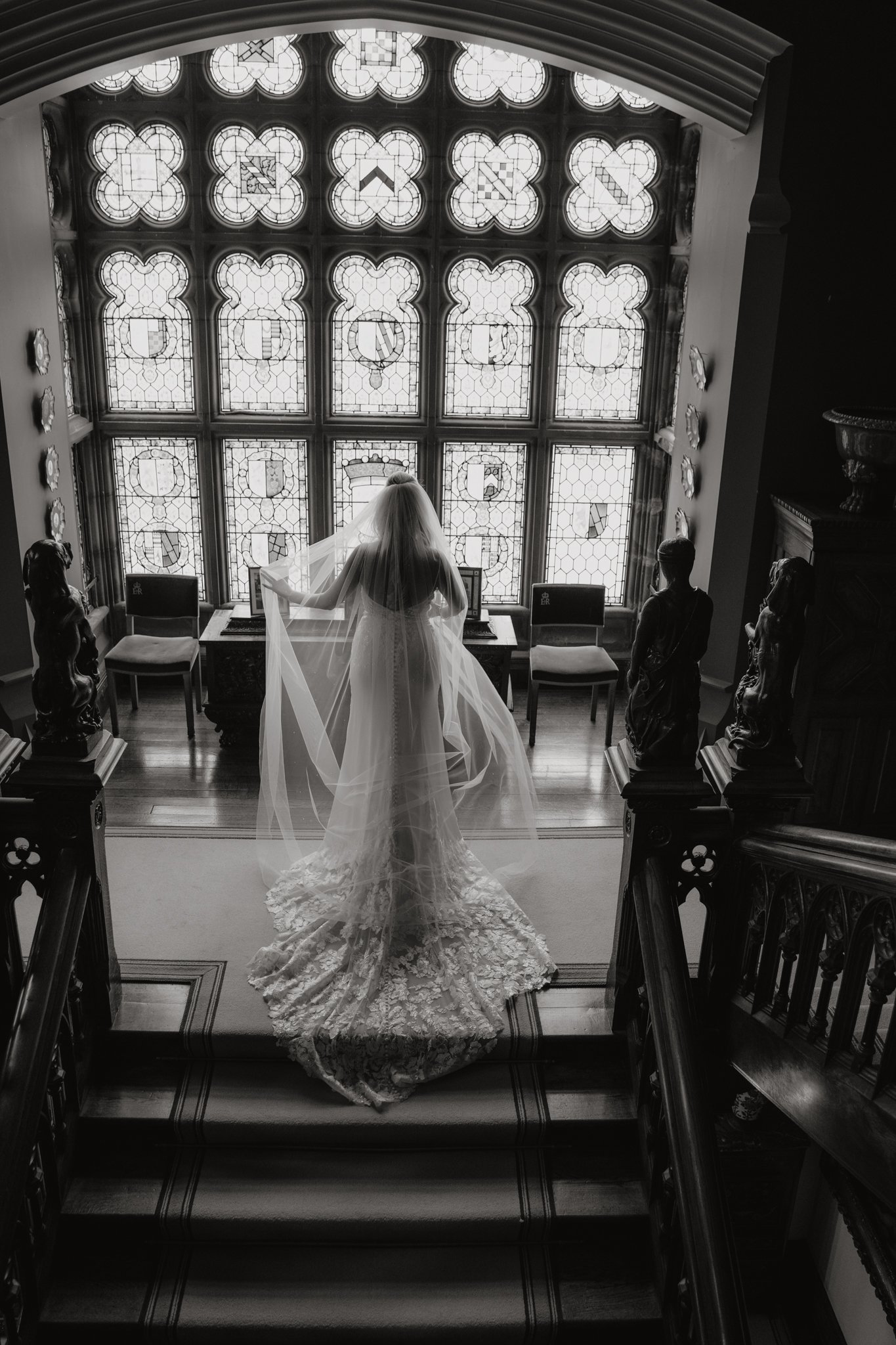 A bride in a wedding dress and veil standing in front of a large stained glass window, holding her veil with her right hand, in a grand interior with ornate wooden railings and statues.