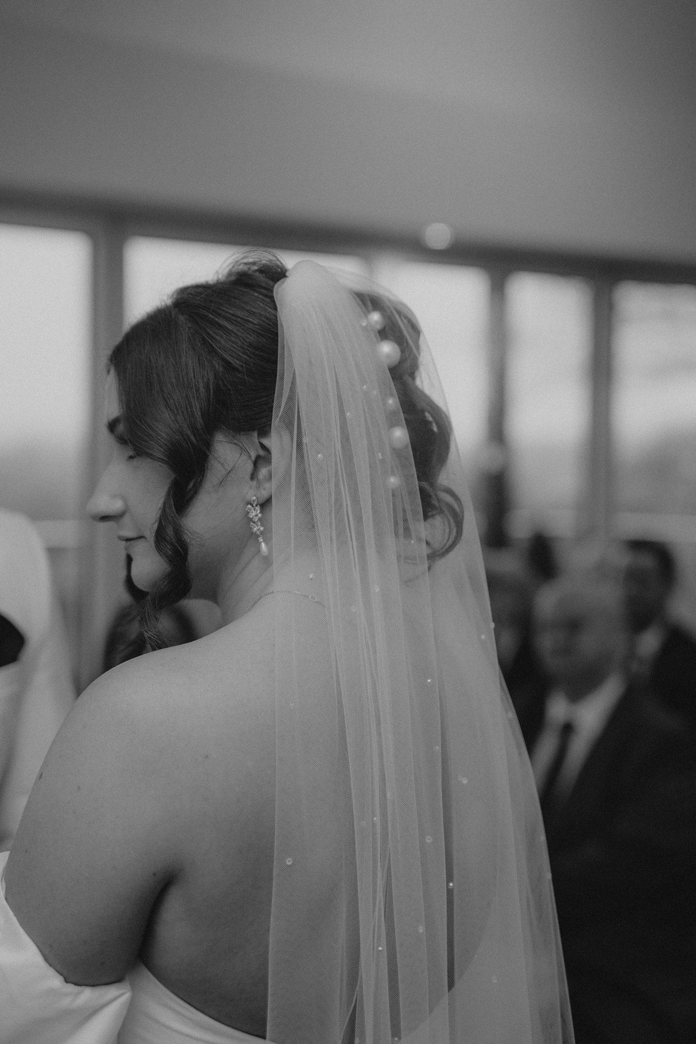 Black and white photo of a bride with dark wavy hair, wearing a veil with pearls, earrings, and a strapless dress, standing in a room with large windows, with some guests visible in the background.