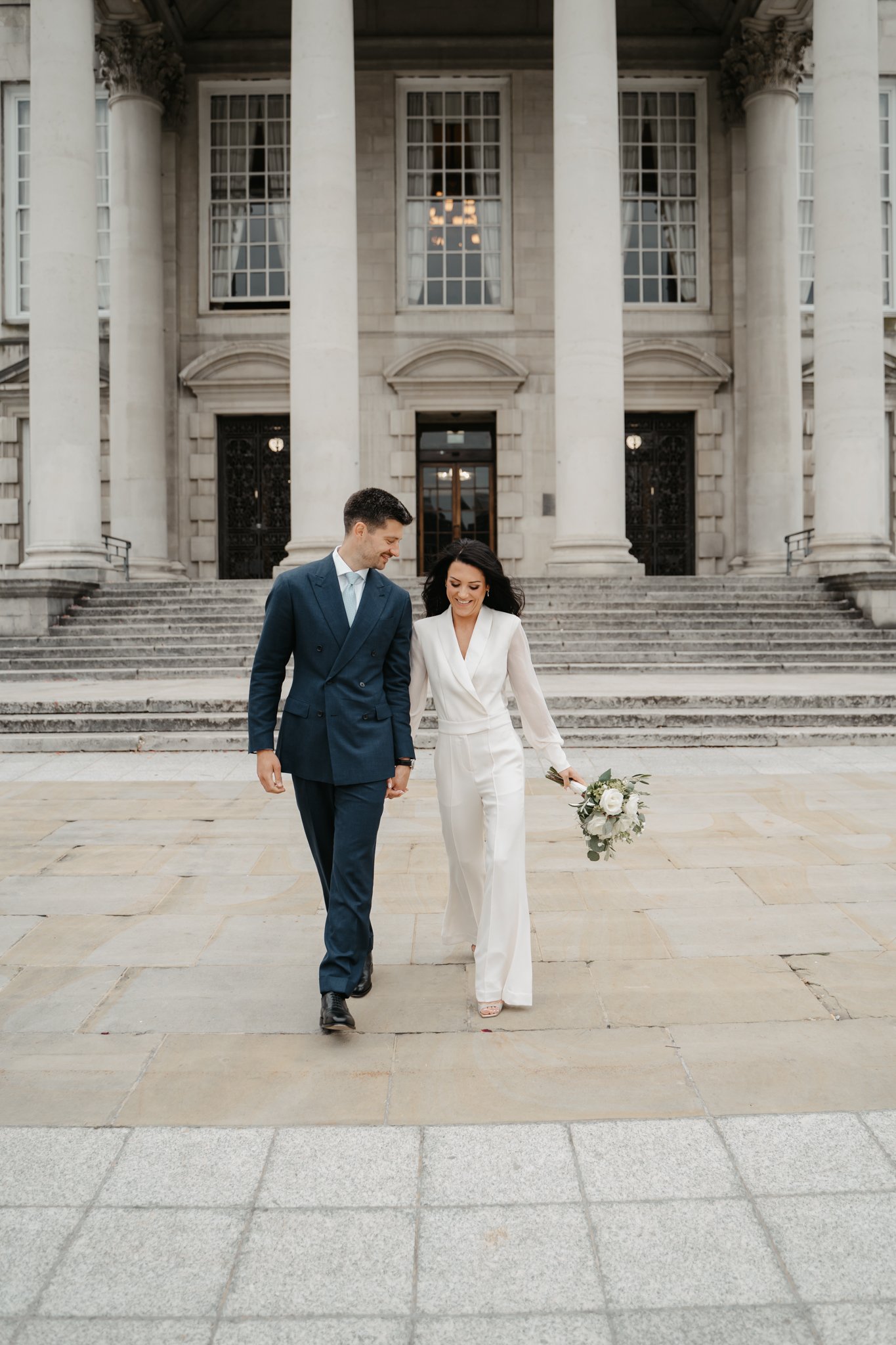 A couple walking hand in hand in front of a grand, historic building with large columns, stairs, and multiple windows. The woman is in a white outfit holding a bouquet, and the man is in a blue suit.