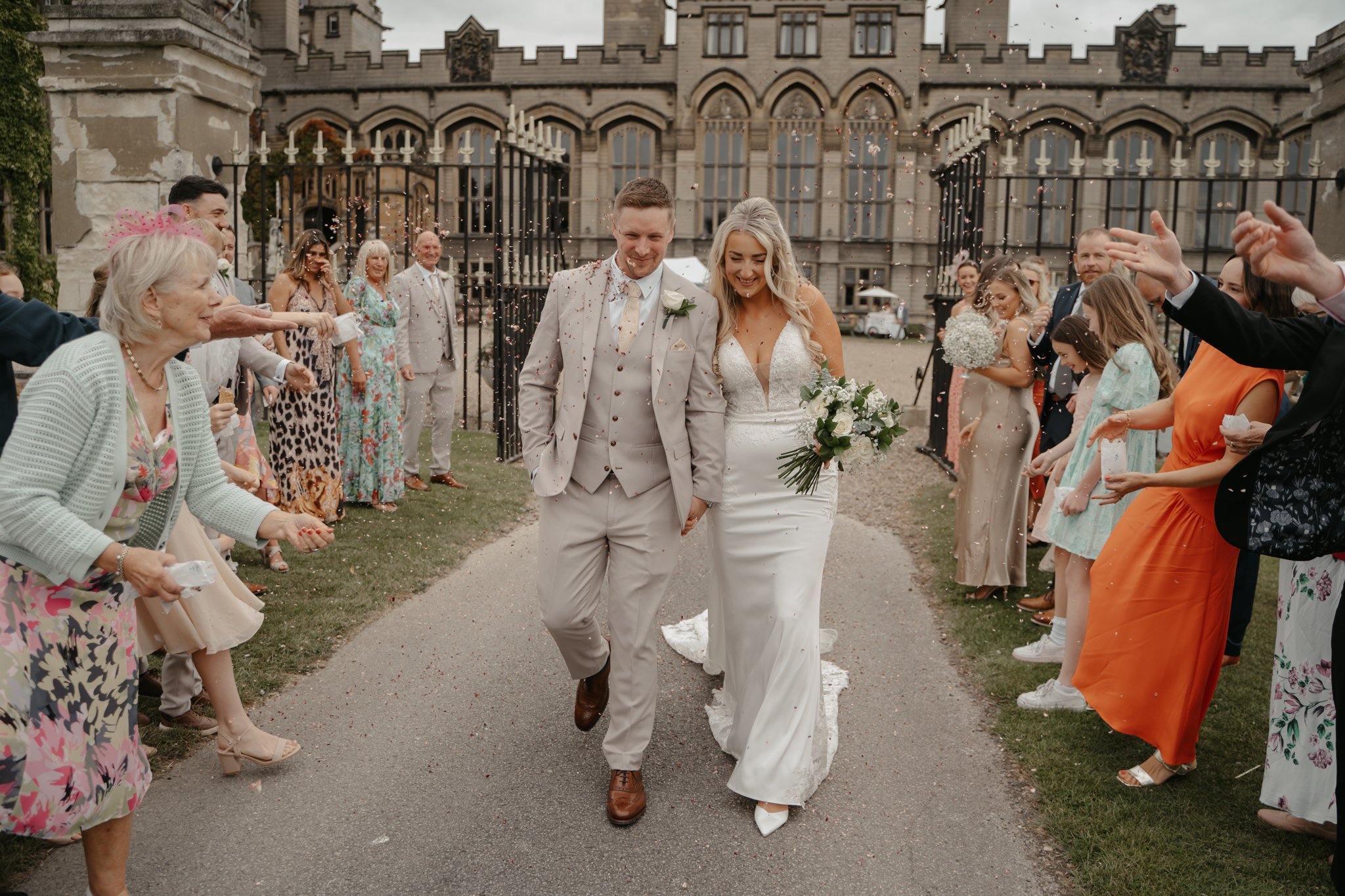 A newlywed couple walking hand-in-hand through a decorated outdoor space surrounded by friends and family celebrating their wedding.