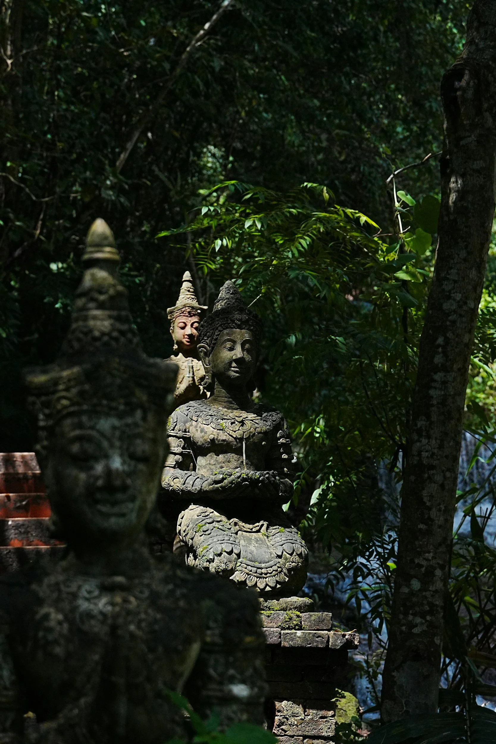 Stone statues of Hindu or Buddhist deities nestled in a lush, green forest, with dappled sunlight filtering through the trees.