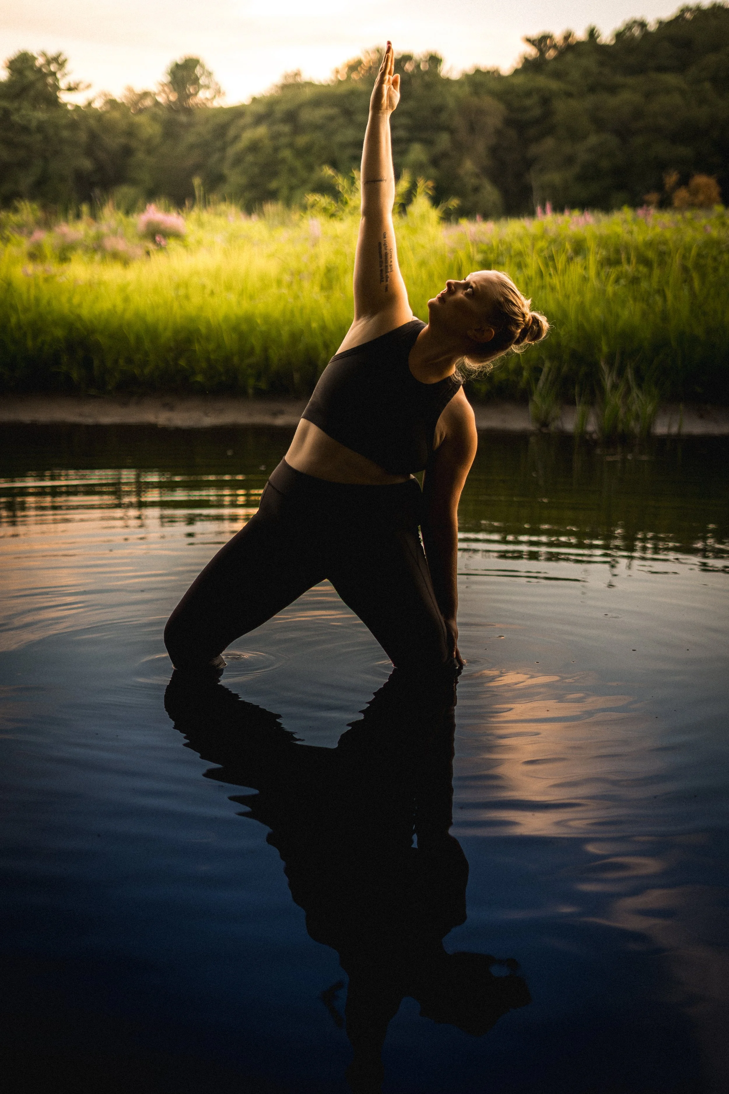 A woman practicing yoga in water at sunset, with her arm raised and standing in a lunge pose. The background shows trees and a soft sunset sky, and her reflection is visible in the water.