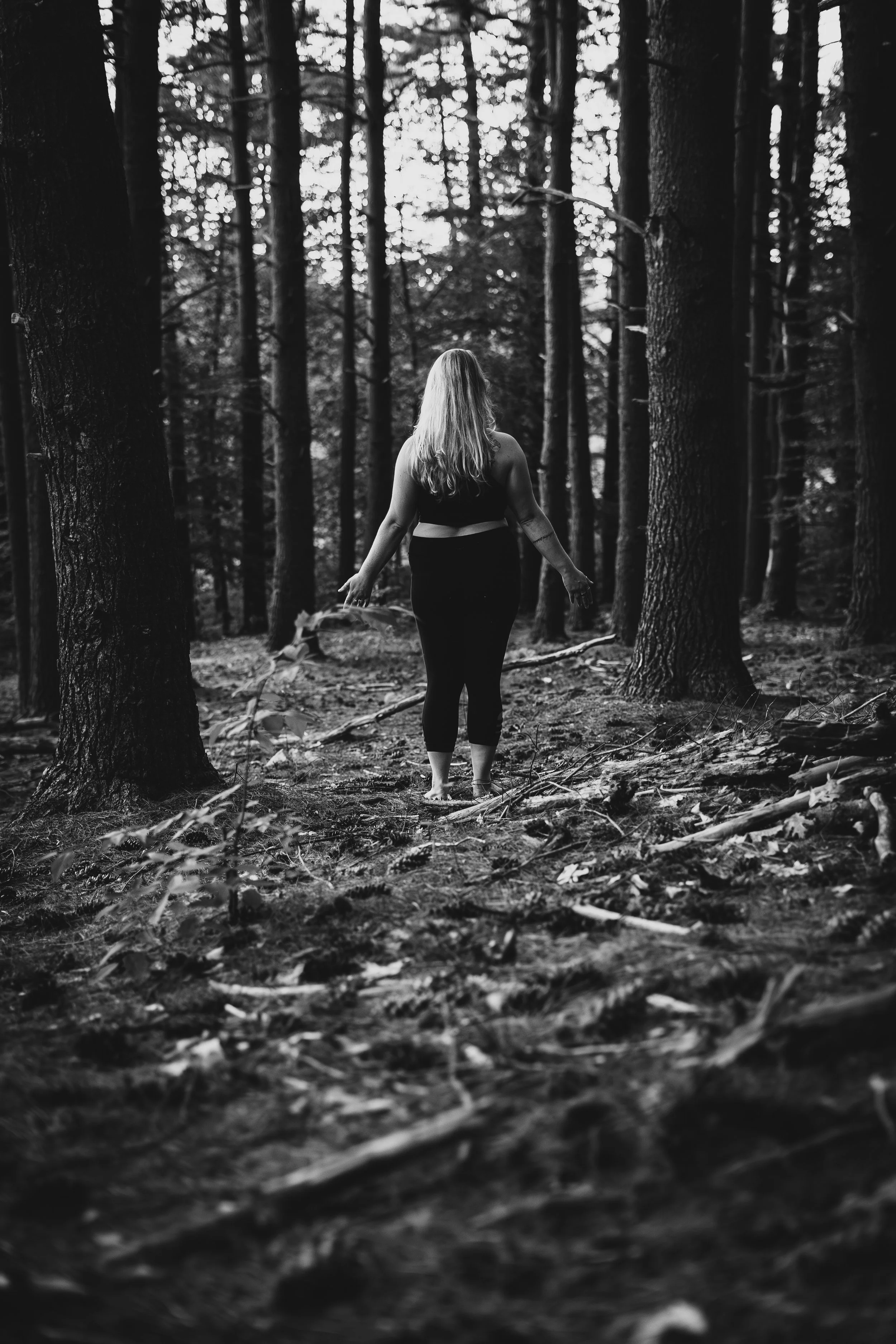 Black and white photo of a woman with long hair walking through a dense forest with tall trees and fallen leaves on the ground.