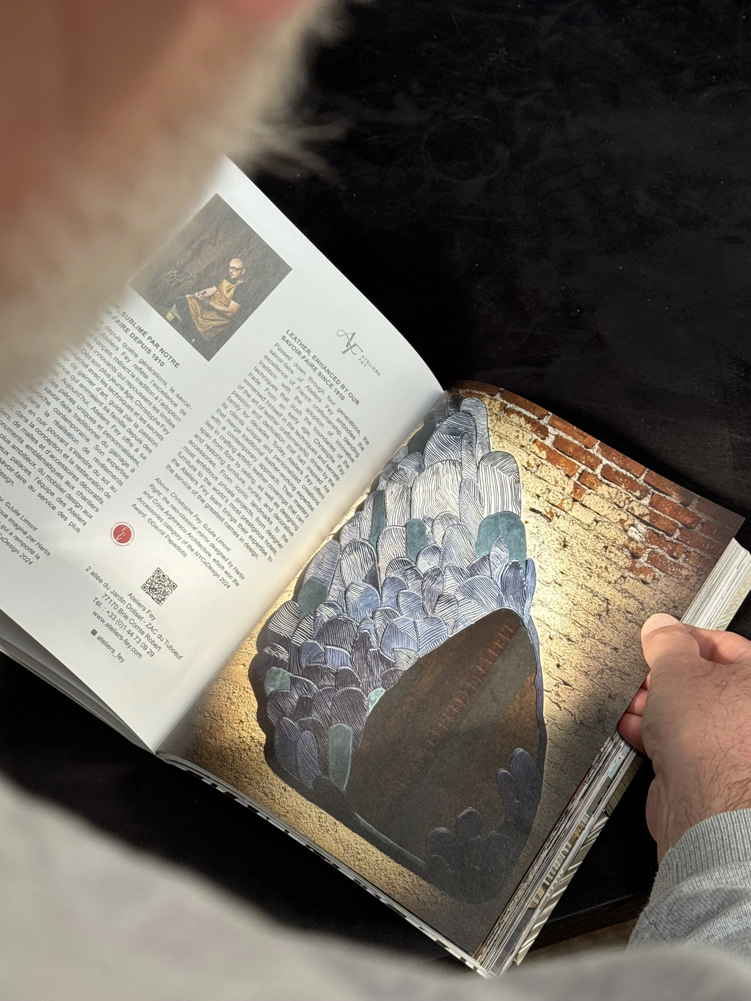 Une personne tient un magazine ouvert, montrant une illustration d'une matière ou sculpture en forme de masse de pierres ou de coquillages en couleurs foncées, posée contre un mur de briques.