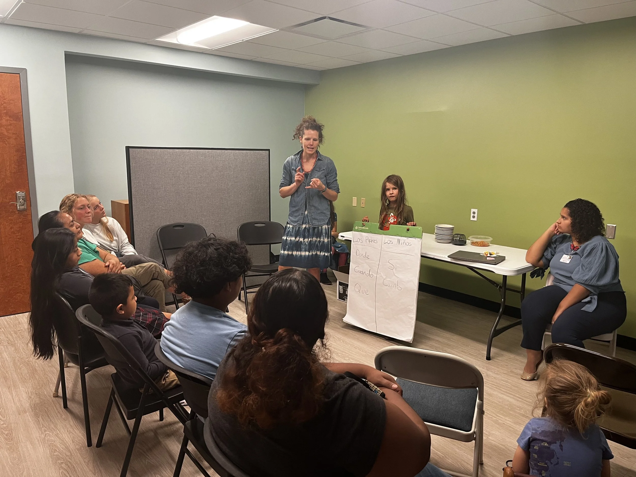 A woman wearing a denim shirt and patterned skirt appears to be giving a presentation to a group of children and adults in a room with green walls. The children are seated, listening, and some are looking at her. There are papers on the table, including a chart titled 'Los Papas' and 'Los Niños' with columns of words in Spanish. The room is meant for a classroom or meeting.