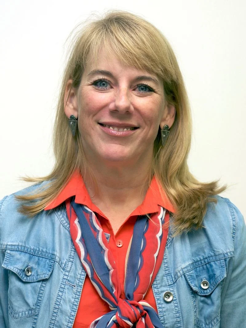 A woman with blonde hair, blue eyes, and light skin, smiling, wearing a denim jacket, red shirt, and a red, white, and blue scarf tied around her neck, with earrings, standing against a plain white background.