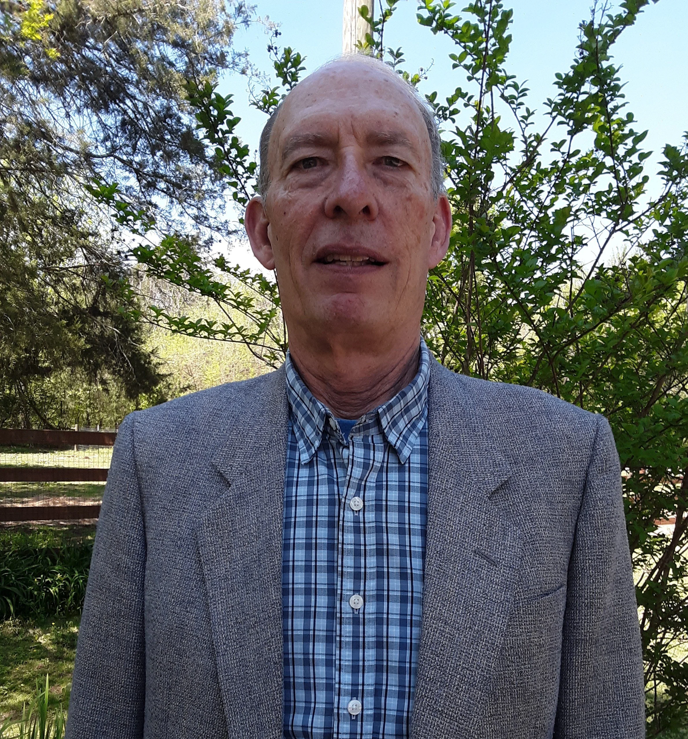 A man with a light complexion and balding head, wearing a gray jacket over a blue checkered shirt, outside in a garden with trees and green leaves in the background.