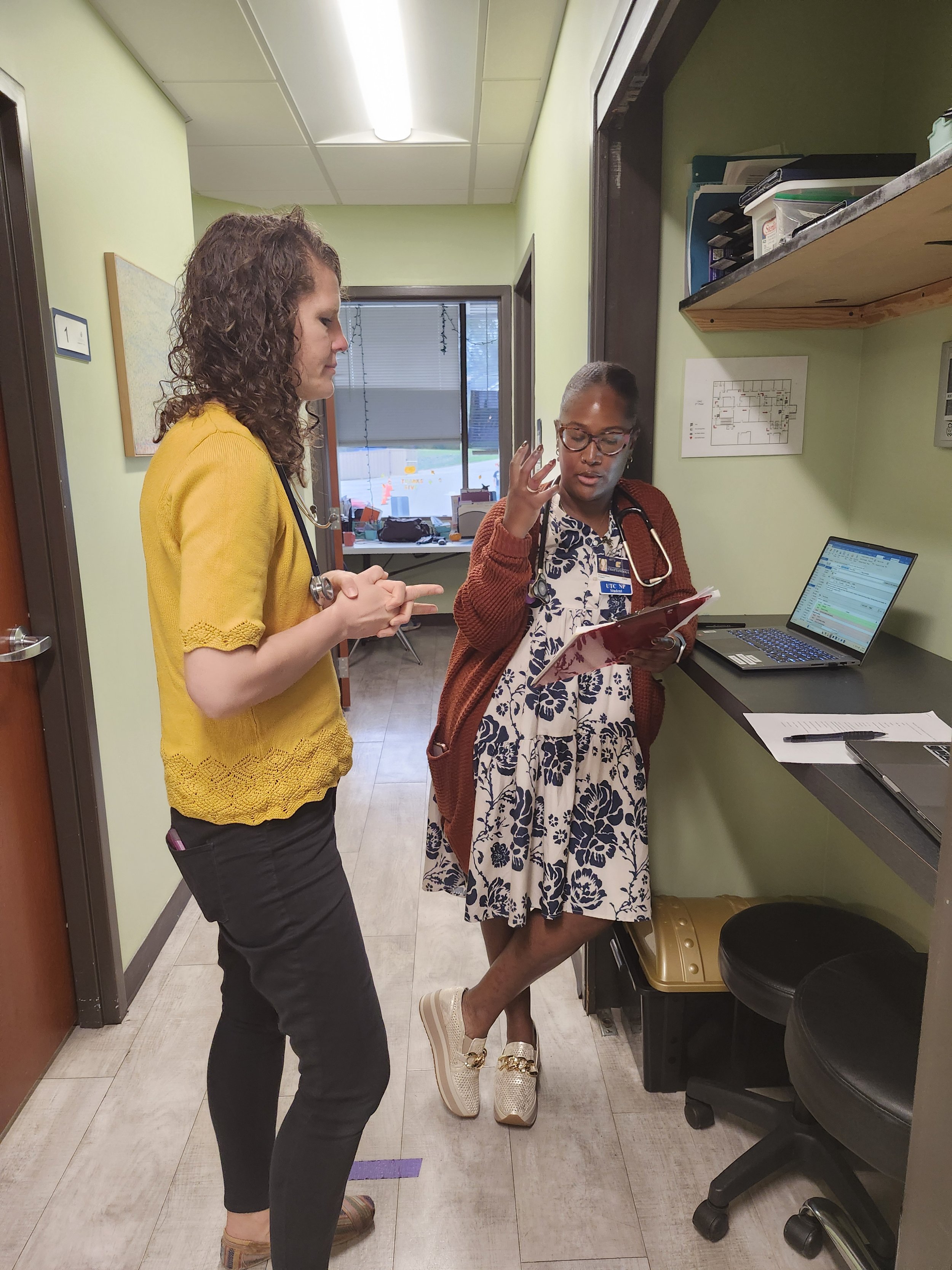 Two women are standing in a medical office, discussing a patient chart. The woman on the left is wearing a yellow top, black pants, and tan shoes. The woman on the right is wearing glasses, a patterned dress, and a rust-colored cardigan, with a stethoscope around her neck. The woman on the right is explaining something while holding the chart, with a laptop on the counter beside her.