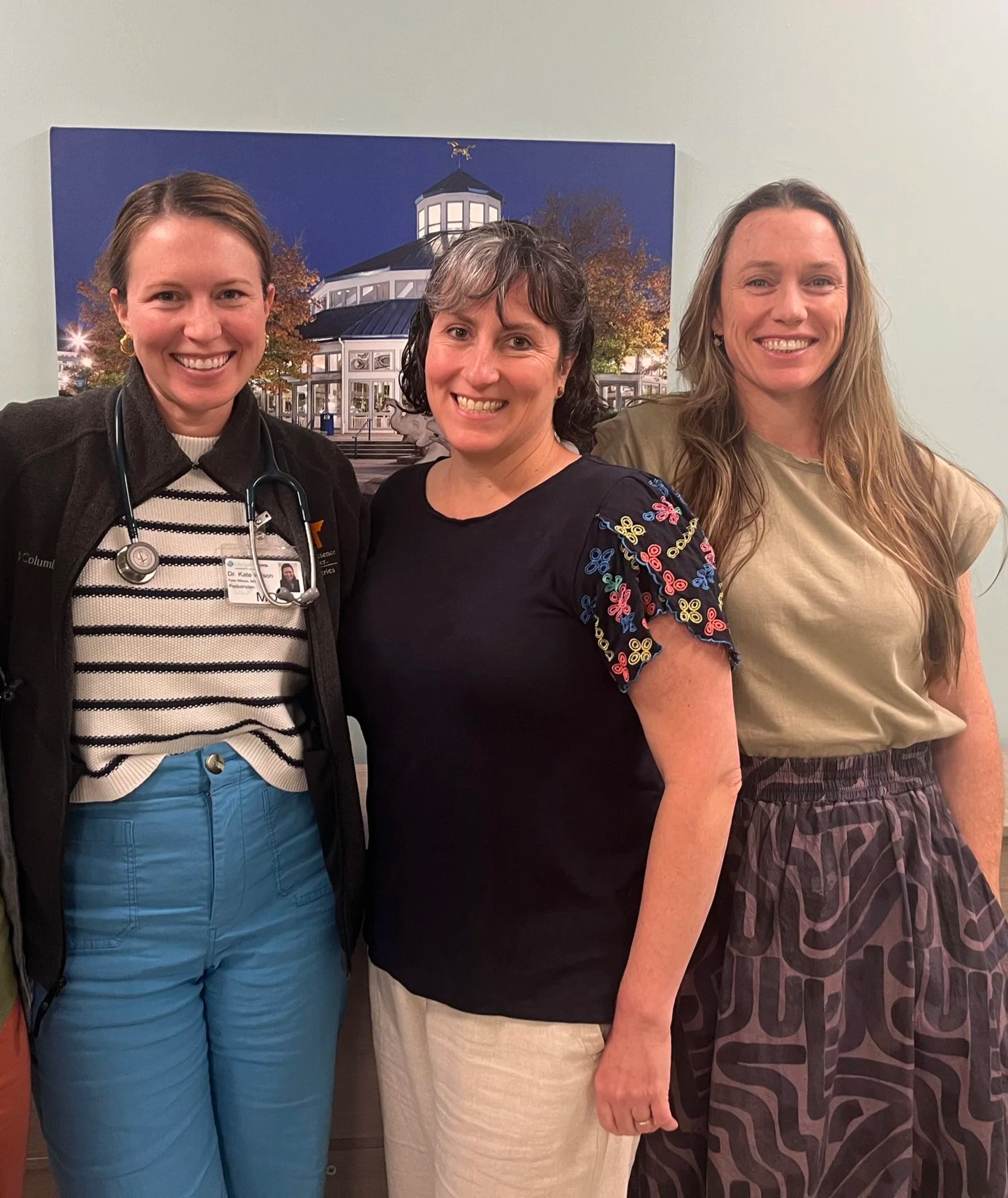 Three women standing together indoors with a large framed picture of a building with a dome and trees in the background behind them. All three women are smiling.