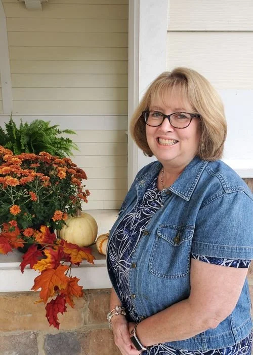 A woman with glasses and short blonde hair smiling while standing outside in front of a window. There are autumn decorations including a pumpkin, red and orange fall leaves, and a potted fern on the windowsill.