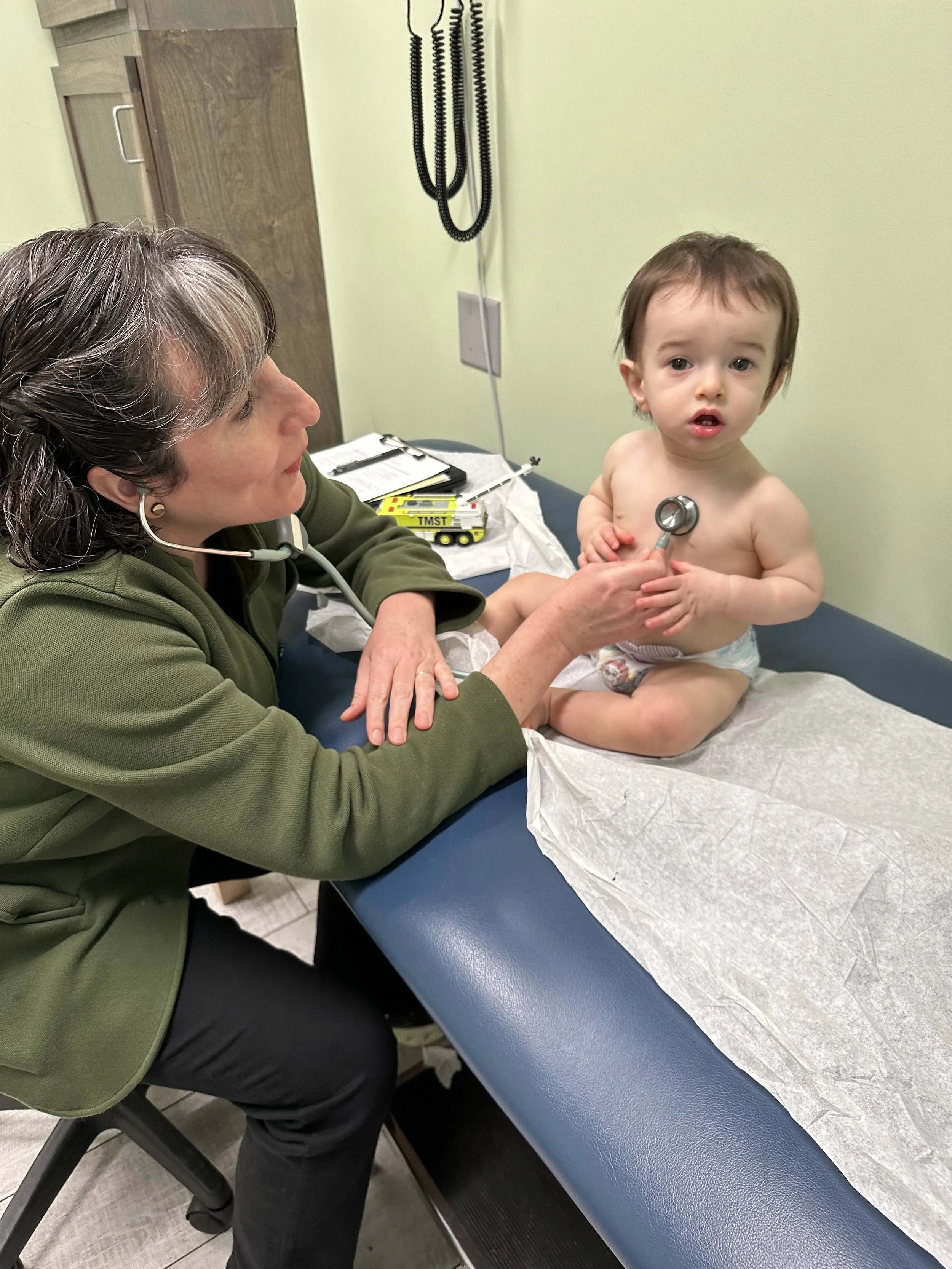 A healthcare professional in a green jacket and black pants performs a medical examination on a young boy with dark hair. The boy is sitting on an examination table with a paper covering, receiving a stethoscope check from the professional. The setting appears to be a doctor's office with medical supplies on a desk nearby.