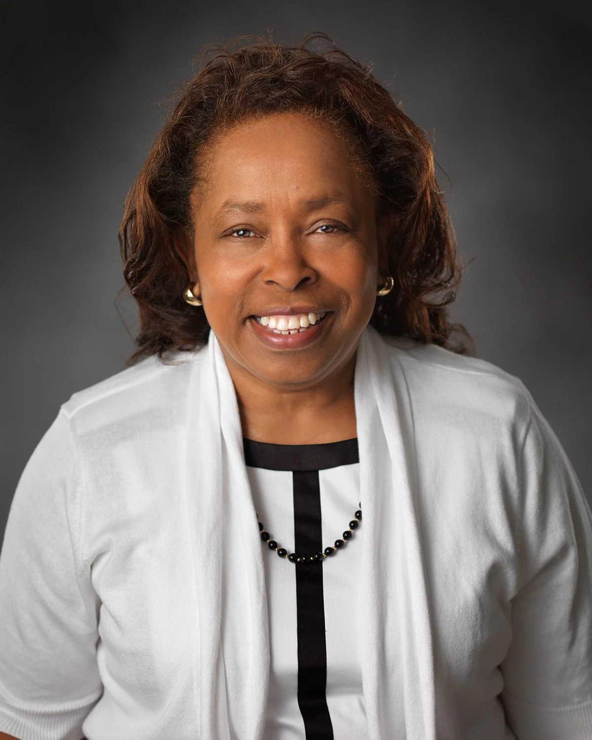 A smiling African American woman with shoulder-length curly hair, wearing a white blazer, black top, and a black necklace, against a dark gray background.