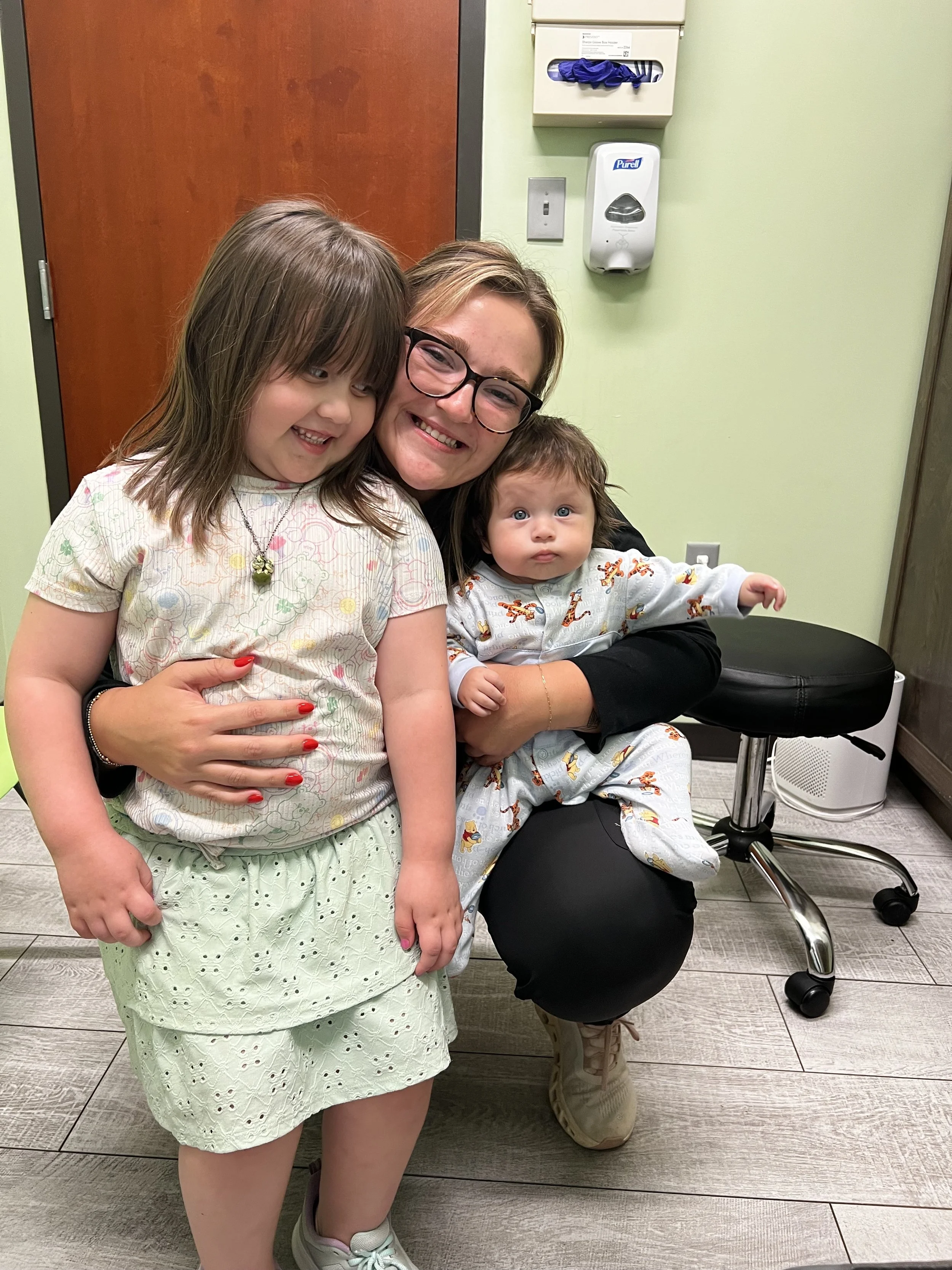 A woman with glasses smiling and two young children in a medical office setting. One child is held in her lap and the other is standing beside her. The room has a wooden door, a wall-mounted hand sanitizer dispenser, tissue box, and medical equipment.