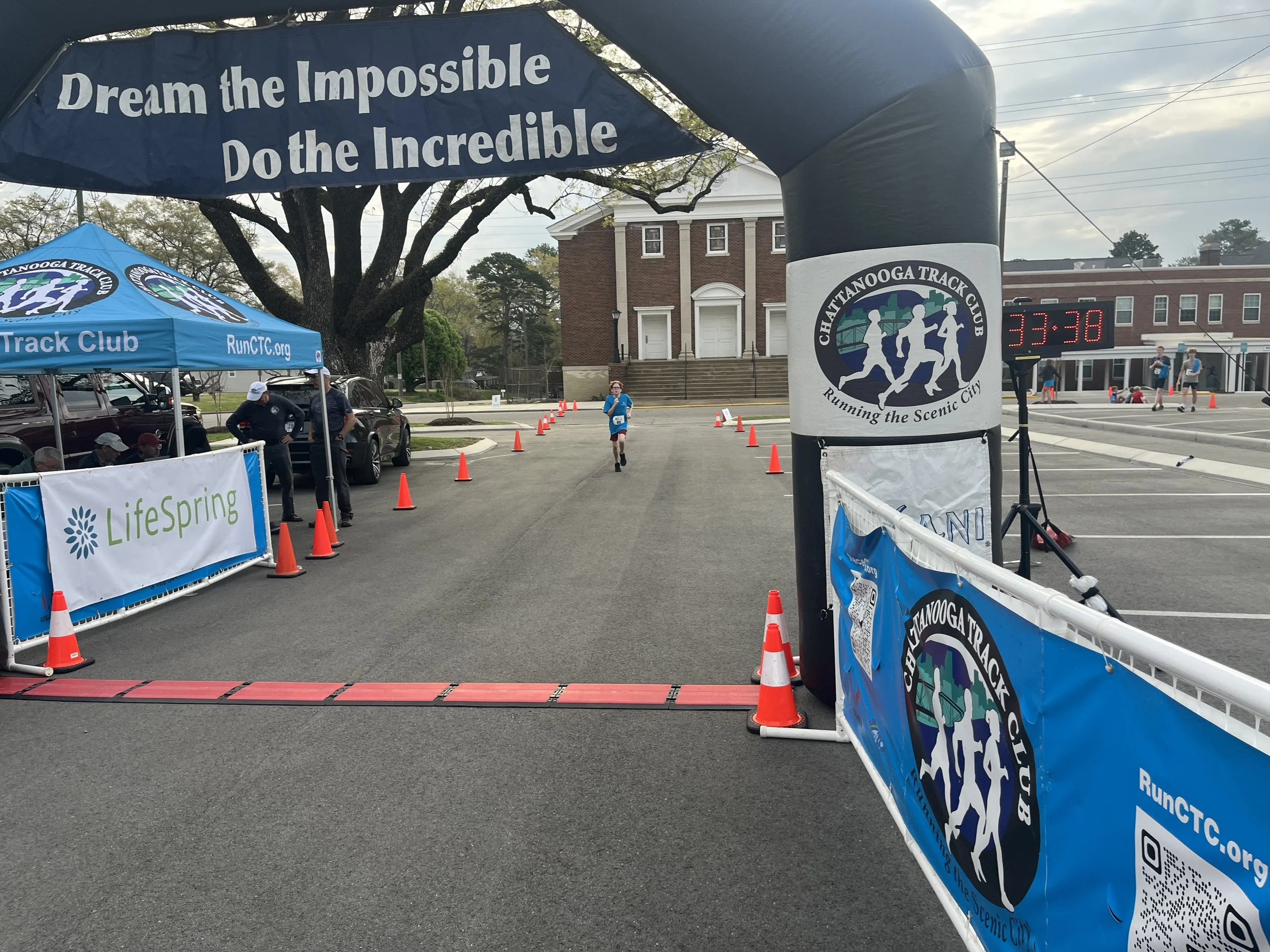 A running race event setup in an outdoor city area with banners and cones. A runner is approaching the finish line, marked by a red mat, with participants and spectators visible in the background. The banners display the Chattanooga Track Club logo and motivational messages.