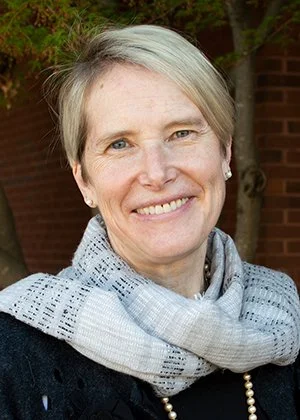 A smiling woman with short blonde hair, wearing a gray scarf, black top, and pearl necklace, outdoors with trees and a brick wall in the background.
