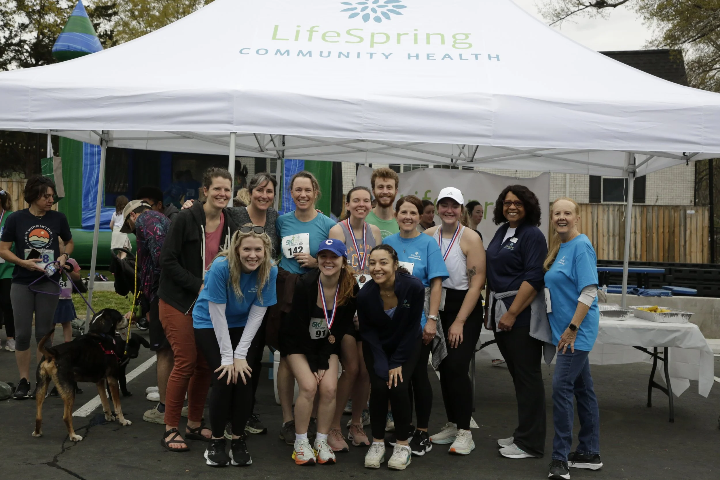 Group of people smiling, some wearing medals, under a UnitySpring Community Health tent at a race event, with a few participants wearing race bibs and medals.