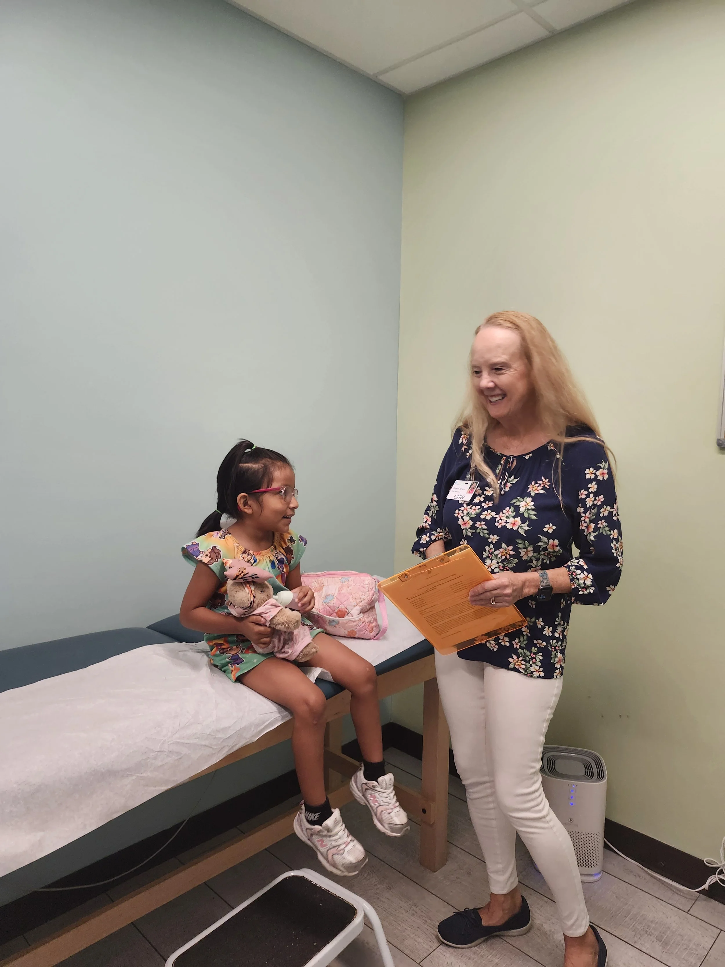 A young girl sitting on an examination table talking to an older woman, likely a healthcare provider, who is standing beside her holding a clipboard. The girl is holding a stuffed animal, and they are inside a medical examination room with light green and beige walls, a stool, and medical equipment nearby.