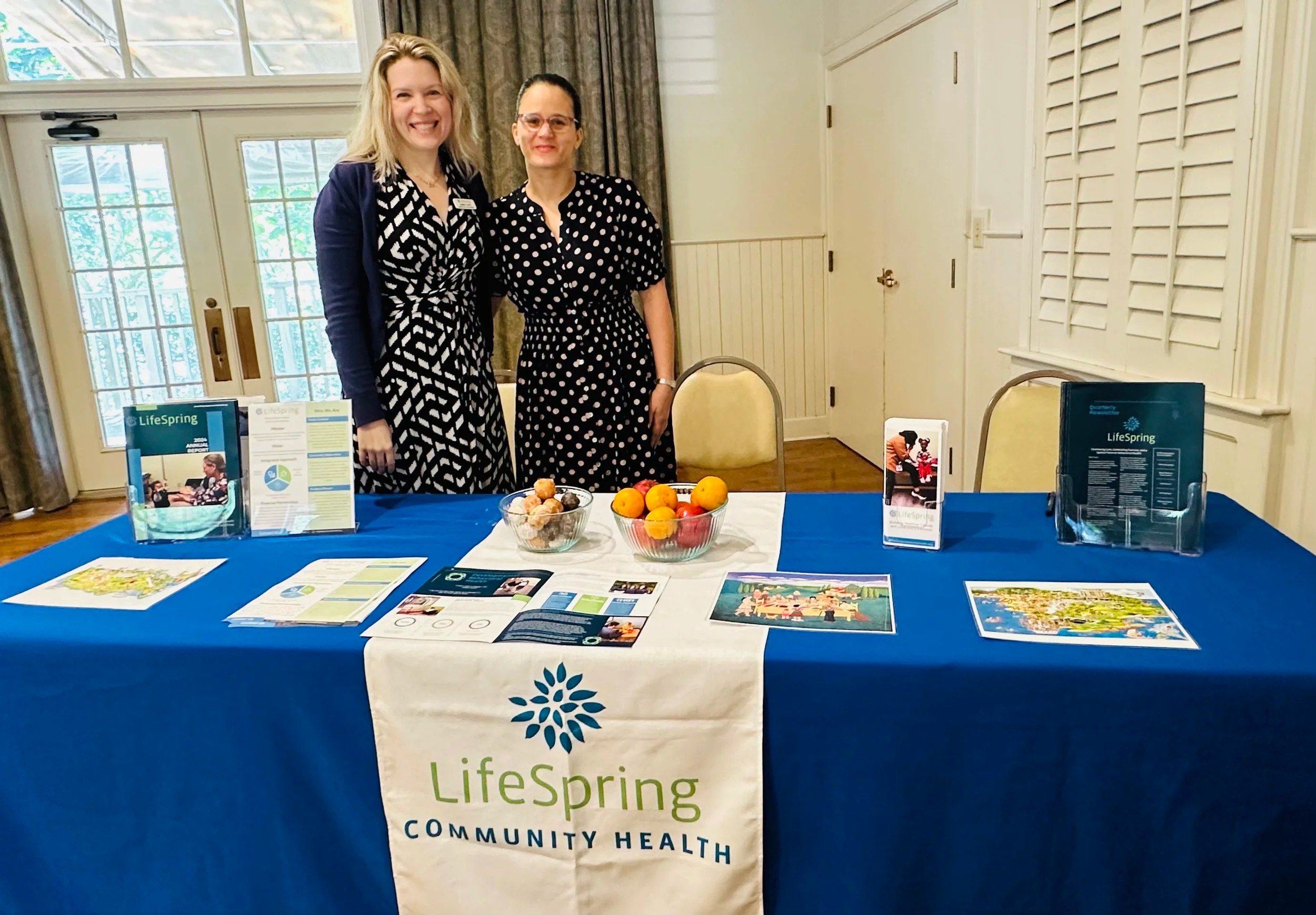 Two women standing behind a table with informational pamphlets and a bowl of peaches, at a Lifespring Community Health event.