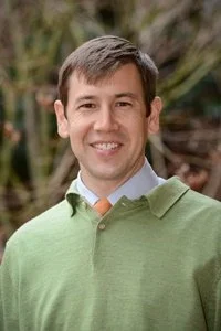 A man smiling outdoors in front of greenery, wearing a light blue shirt and a green sweater.
