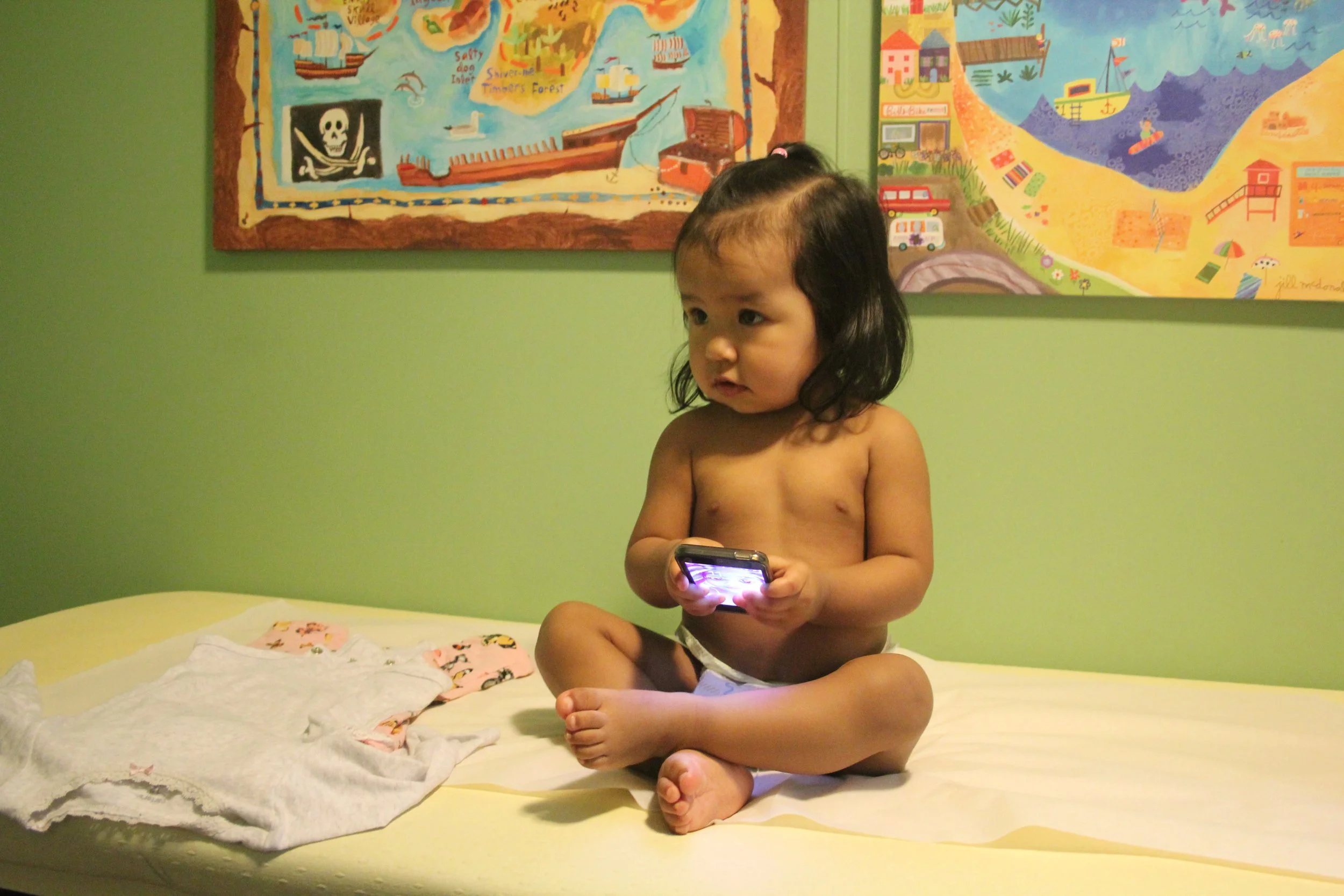 A young child with dark hair, sitting on a bed, looking at a phone glowing in their hands. The child is shirtless and has a calm expression. The bed has white and pink patterned bedding. In the background, there are colorful children's drawings on a green wall.