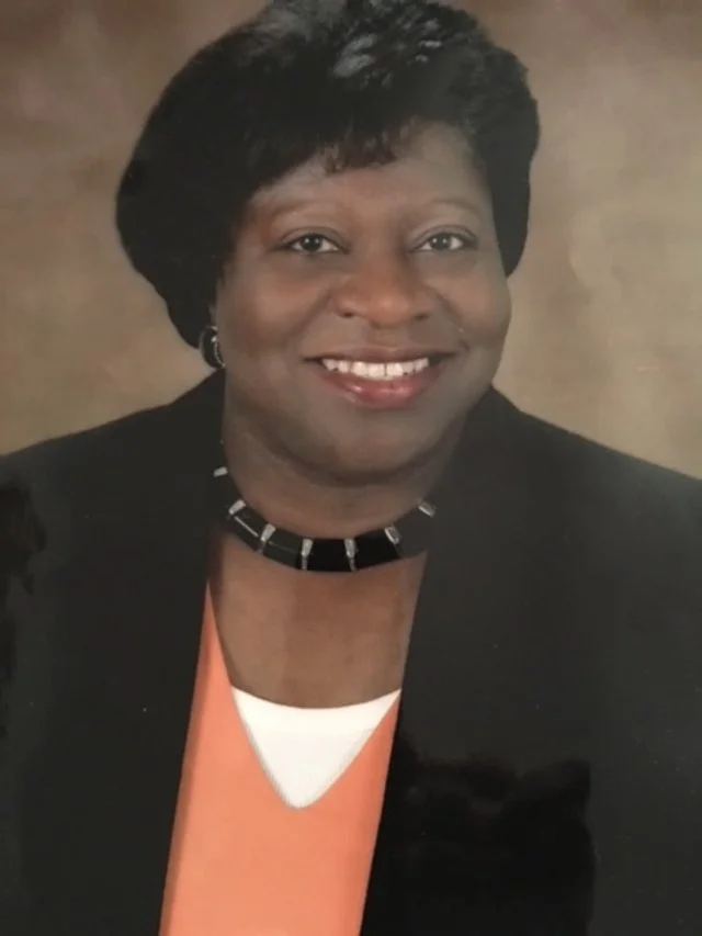 A smiling African American woman with short black hair, wearing a black blazer, an orange top, a white undershirt, and a black necklace with silver accents.