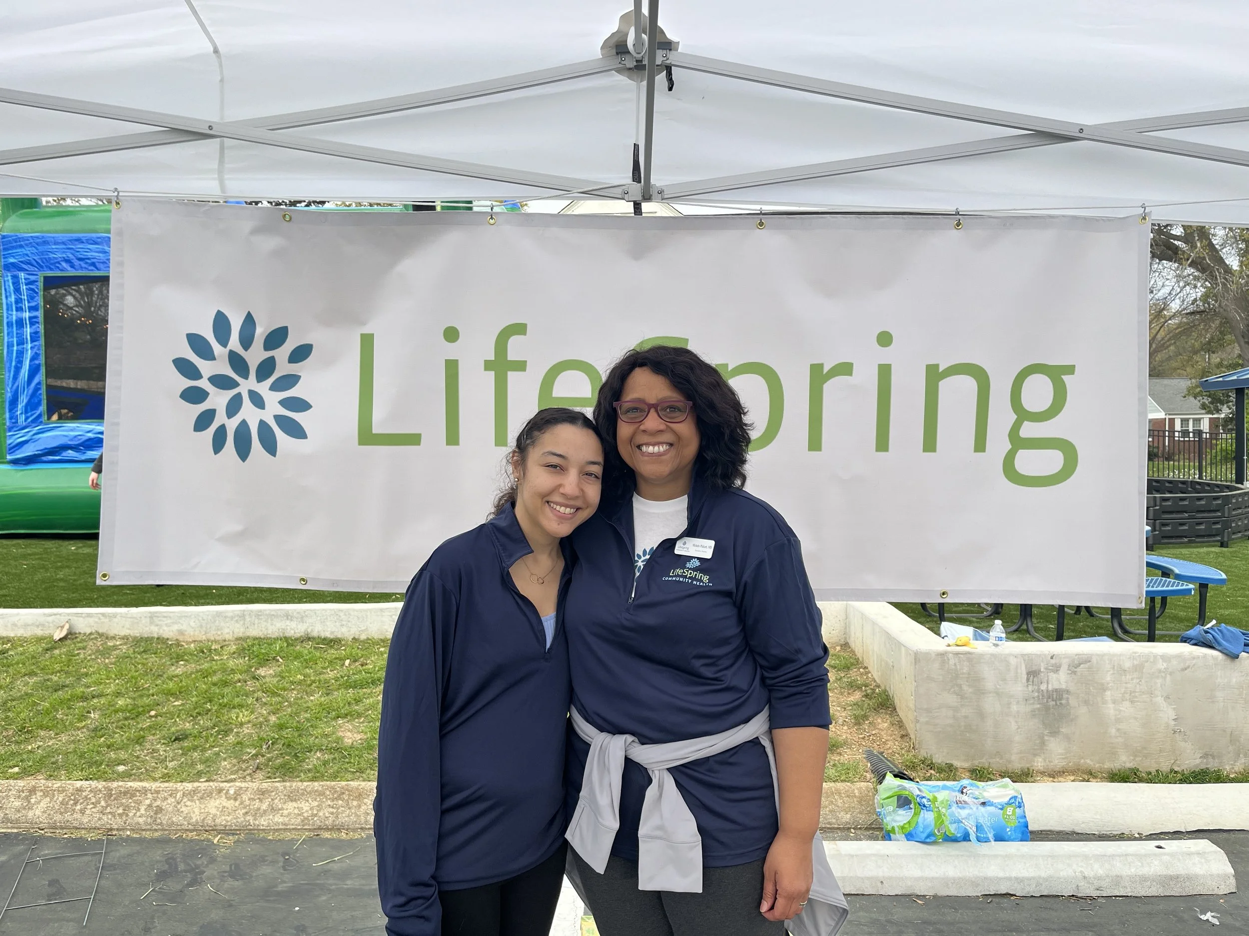 Two women standing under a white canopy tent at an outdoor event, smiling. Behind them, a large banner with the logo and text 'Lifespring' is visible. One woman wears glasses and a navy jacket, the other has her hair tied back and wears a navy jacket tied around her waist.