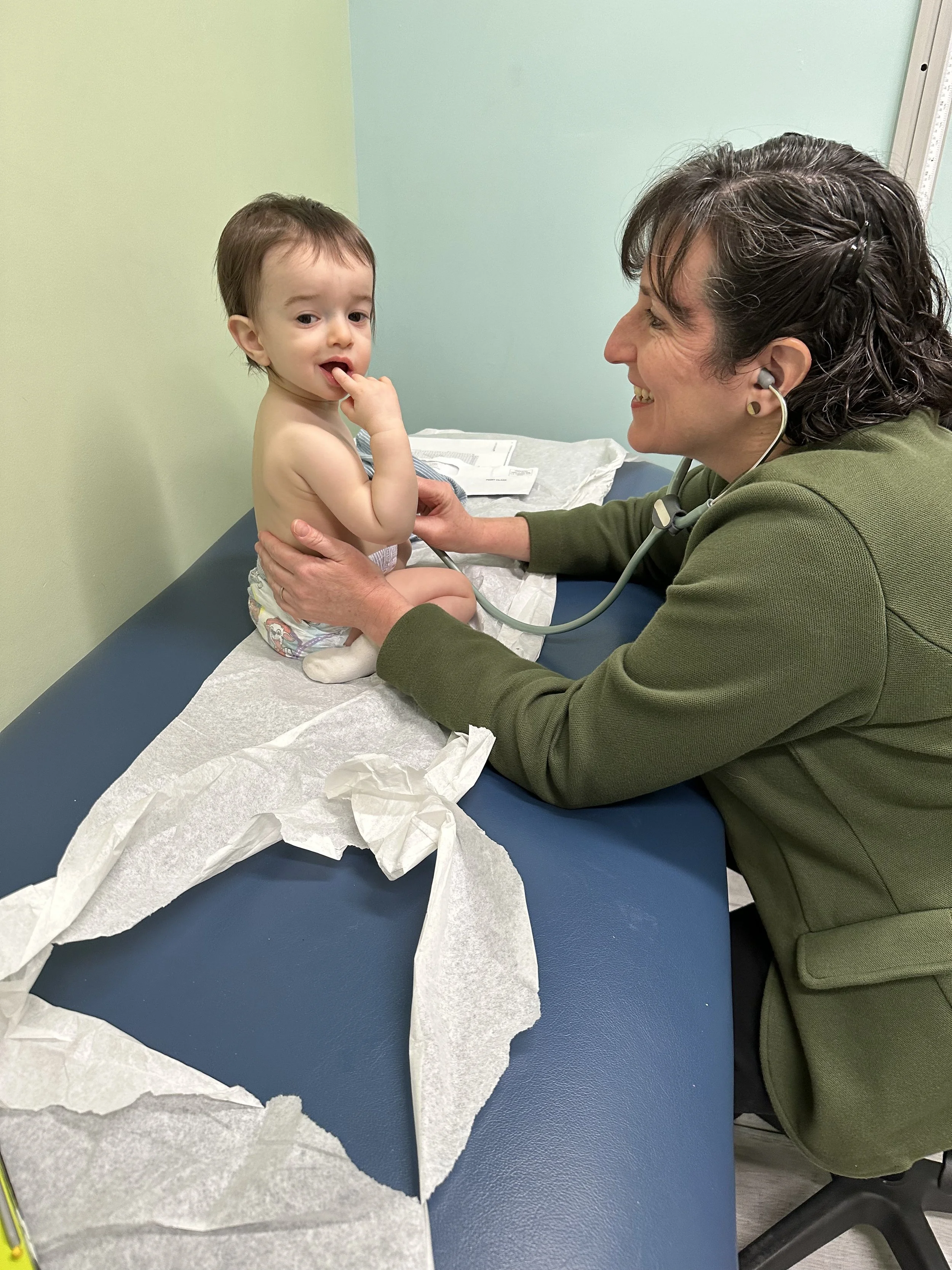 A healthcare professional, smiling, using a stethoscope to examine a young boy sitting on an examination table. The boy is shirtless, wearing only a diaper, with his finger near his mouth. There are medical supplies and tissue paper on the table.