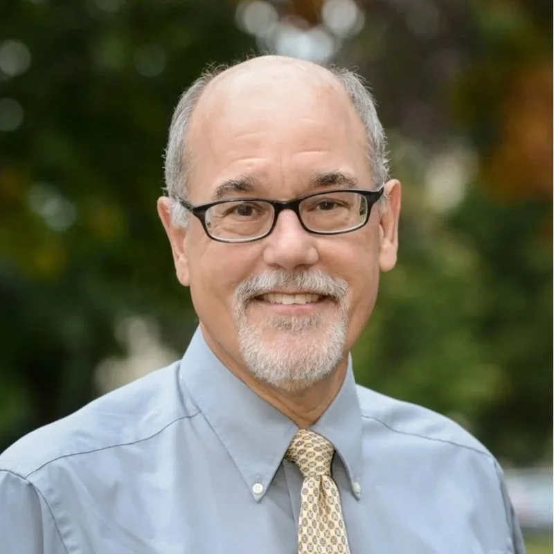 Portrait of a middle-aged man with glasses, a goatee, and a friendly smile, dressed in a light blue shirt and patterned tie, outdoors with blurred greenery in the background.