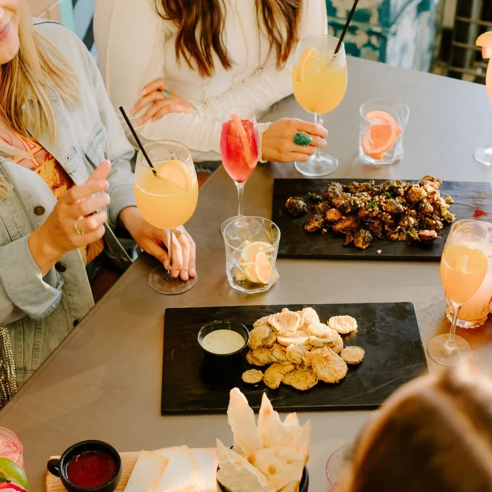 A group of people sitting around a table with drinks and dishes of fried food, including chicken and vegetables, at a restaurant or cafe.