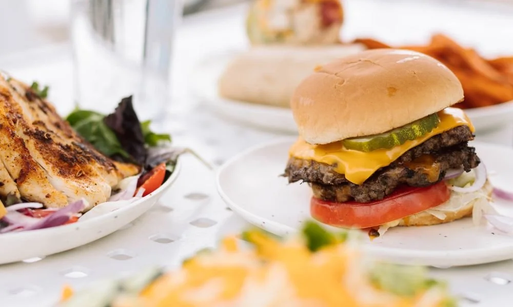 Close-up of a cheeseburger with pickles, tomato, lettuce, melted cheese, and a beef patty on a sandwich bun, with sides of grilled chicken and sweet potato fries on a table.