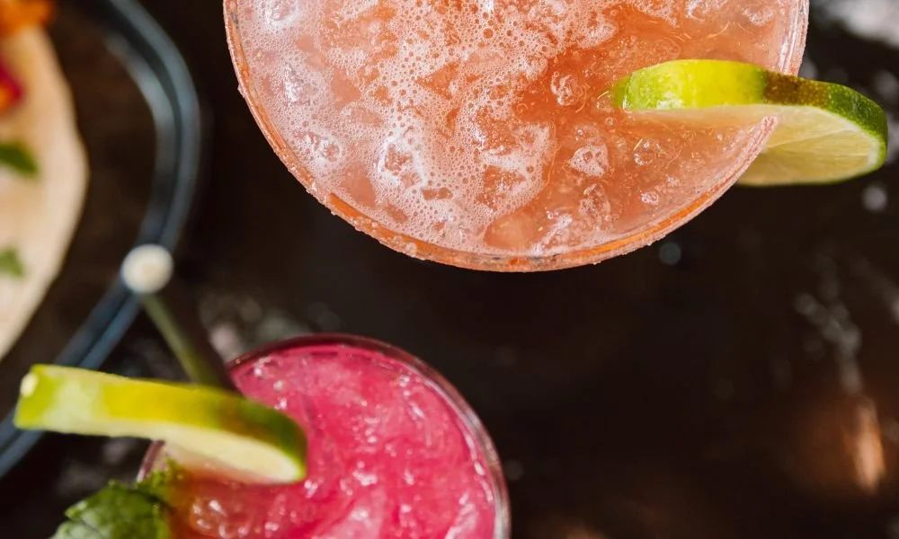 Two colorful cocktails with lime wedges on a dark surface.