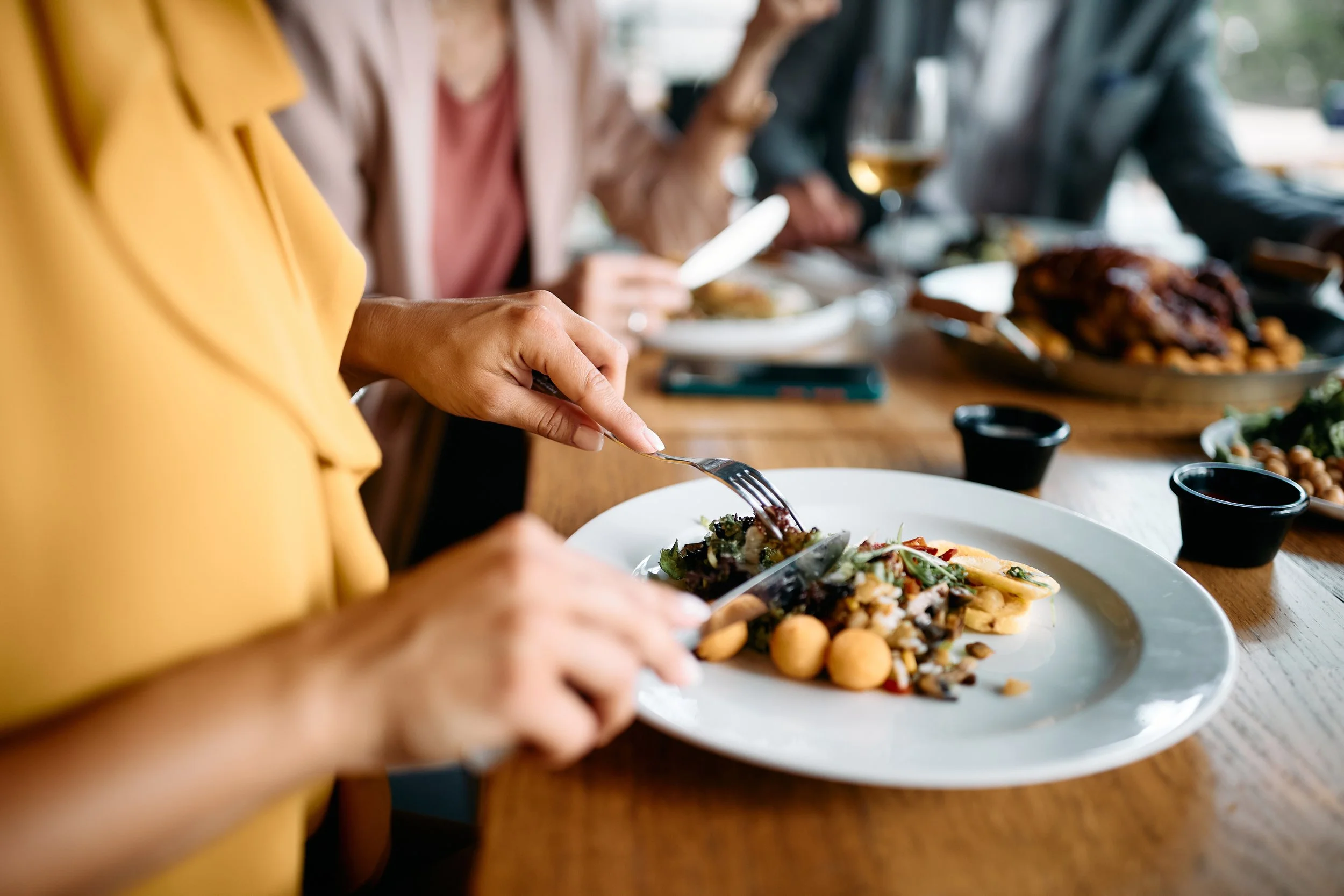 People dining at a restaurant, with focus on a person in a yellow shirt cutting a salad on a white plate. The table has various dishes, drinks, and condiments.