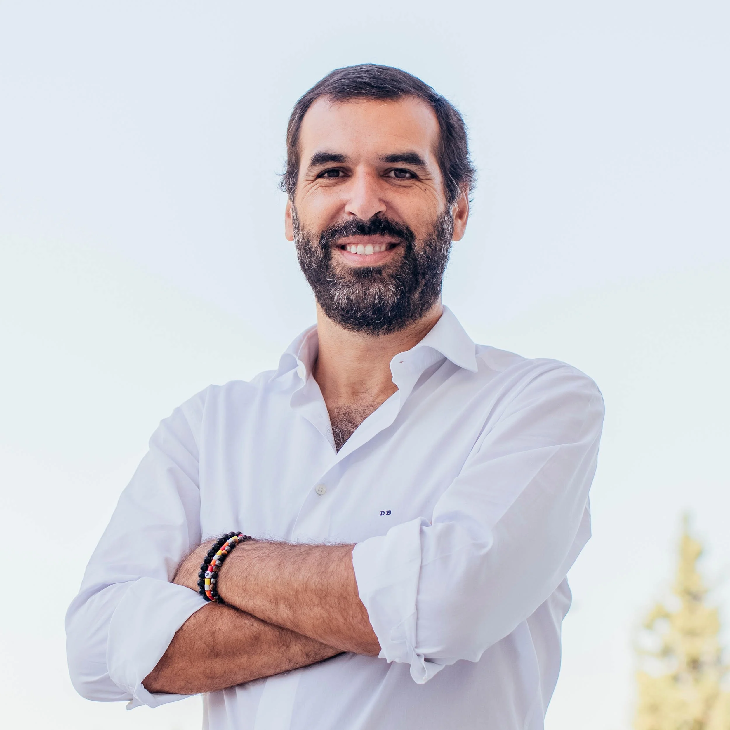 A man with dark hair and a beard smiling with arms crossed, wearing a white button-up shirt, outdoors with a clear sky in the background.