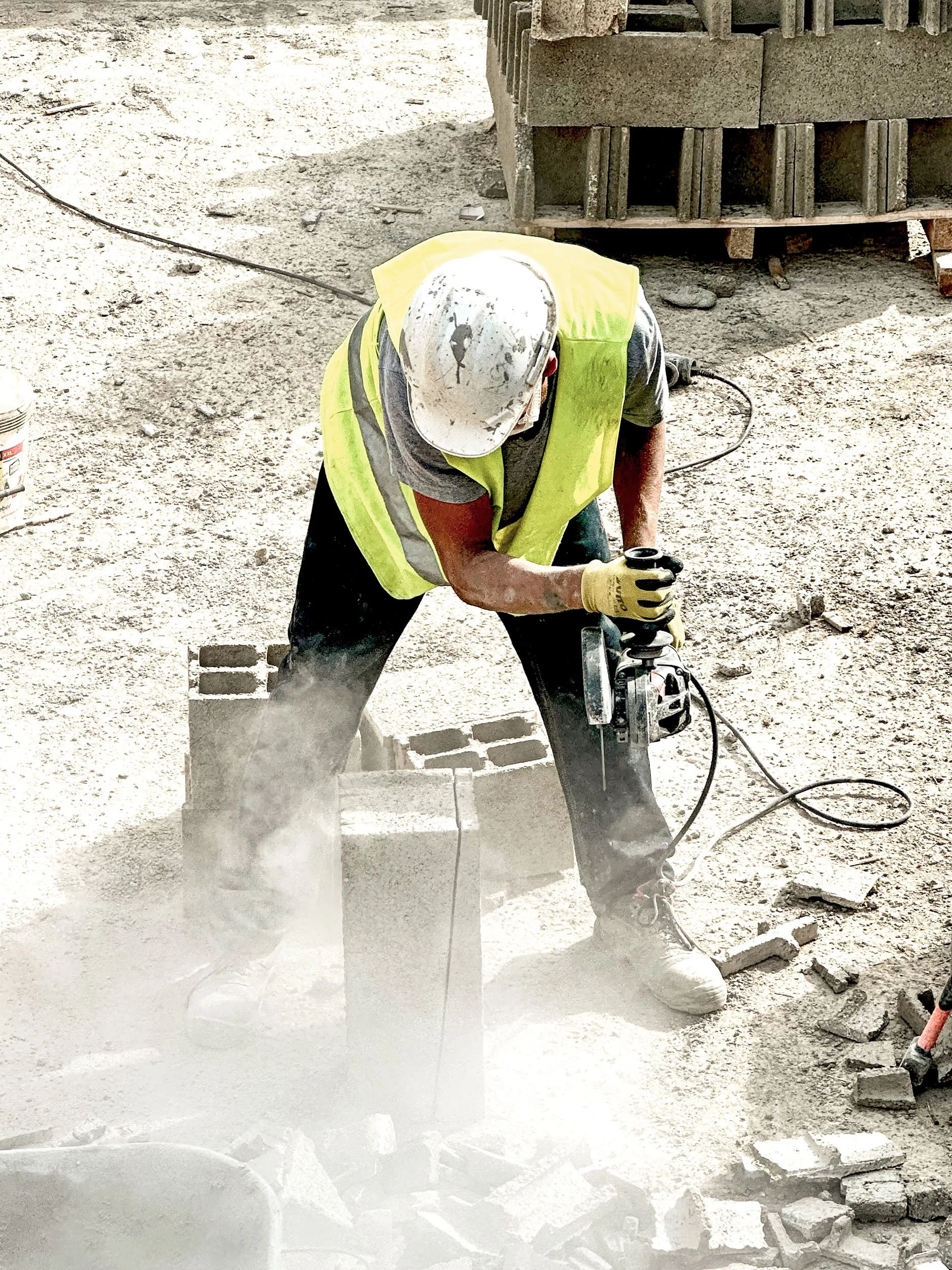 A construction worker wearing a white hard hat, yellow safety vest, and gloves using a power tool to cut bricks on a construction site.