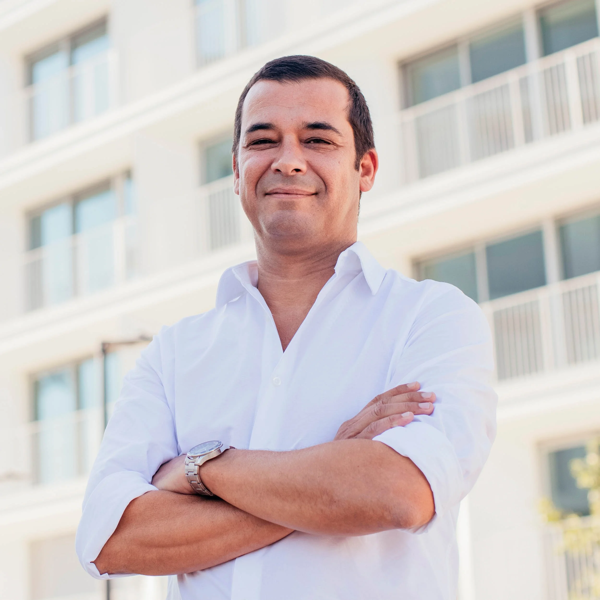Image of a confident man with crossed arms smiling, standing in front of a modern apartment building, wearing a white shirt and a wristwatch.