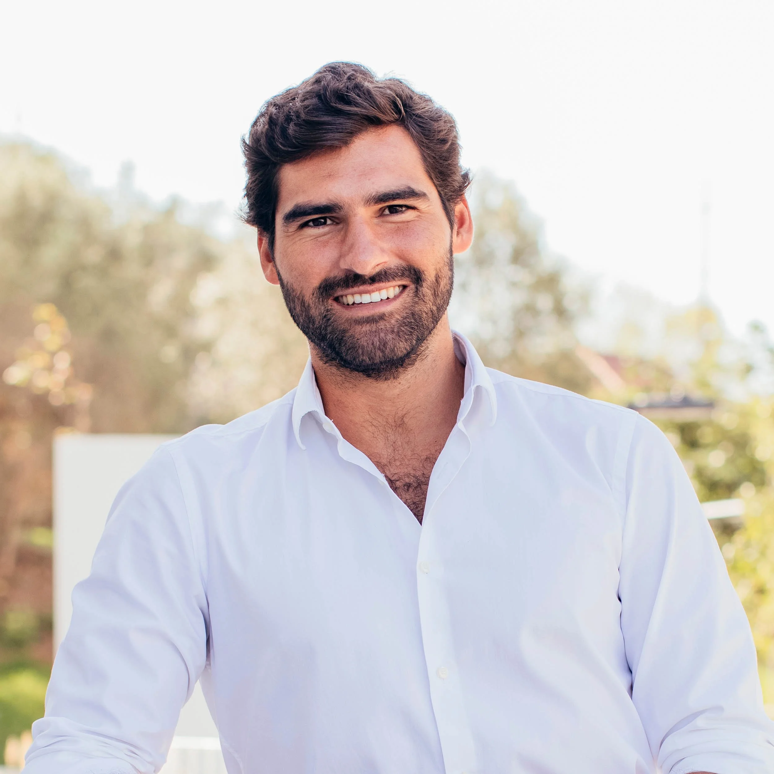 A smiling man with dark, wavy hair and a beard, wearing a white button-down shirt, outdoors with blurred trees in the background.