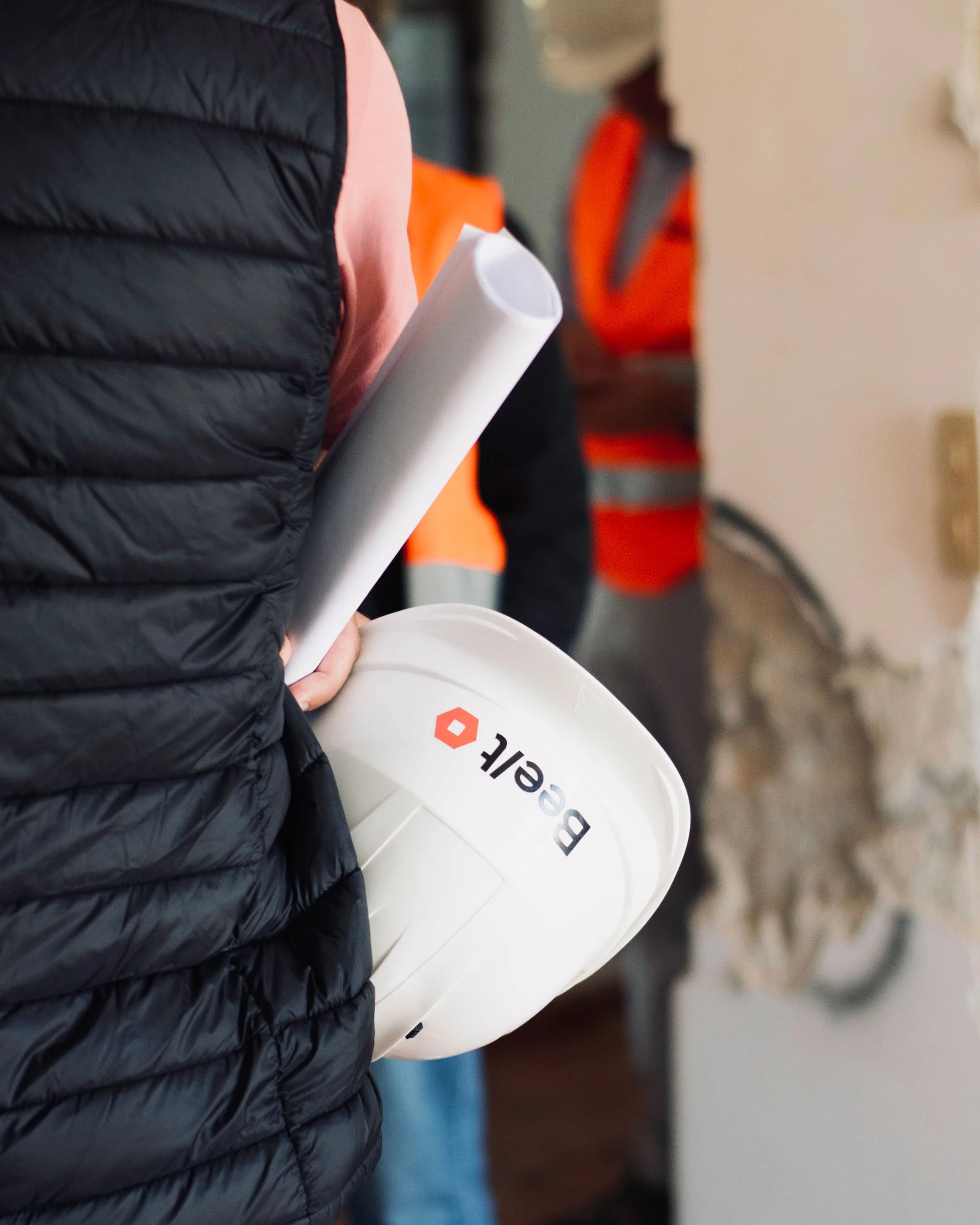 Person holding a rolled-up blueprint and a white hard hat with 'BECL' logo, standing at a construction site with safety vests in the background.
