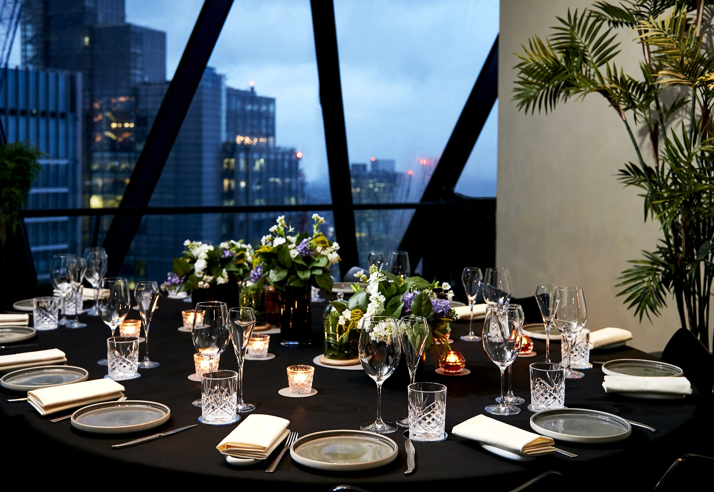 A dinner table set for a formal event in a high-rise restaurant with city skyline views, featuring floral centerpieces, candles, glassware, plates, napkins, and cutlery.