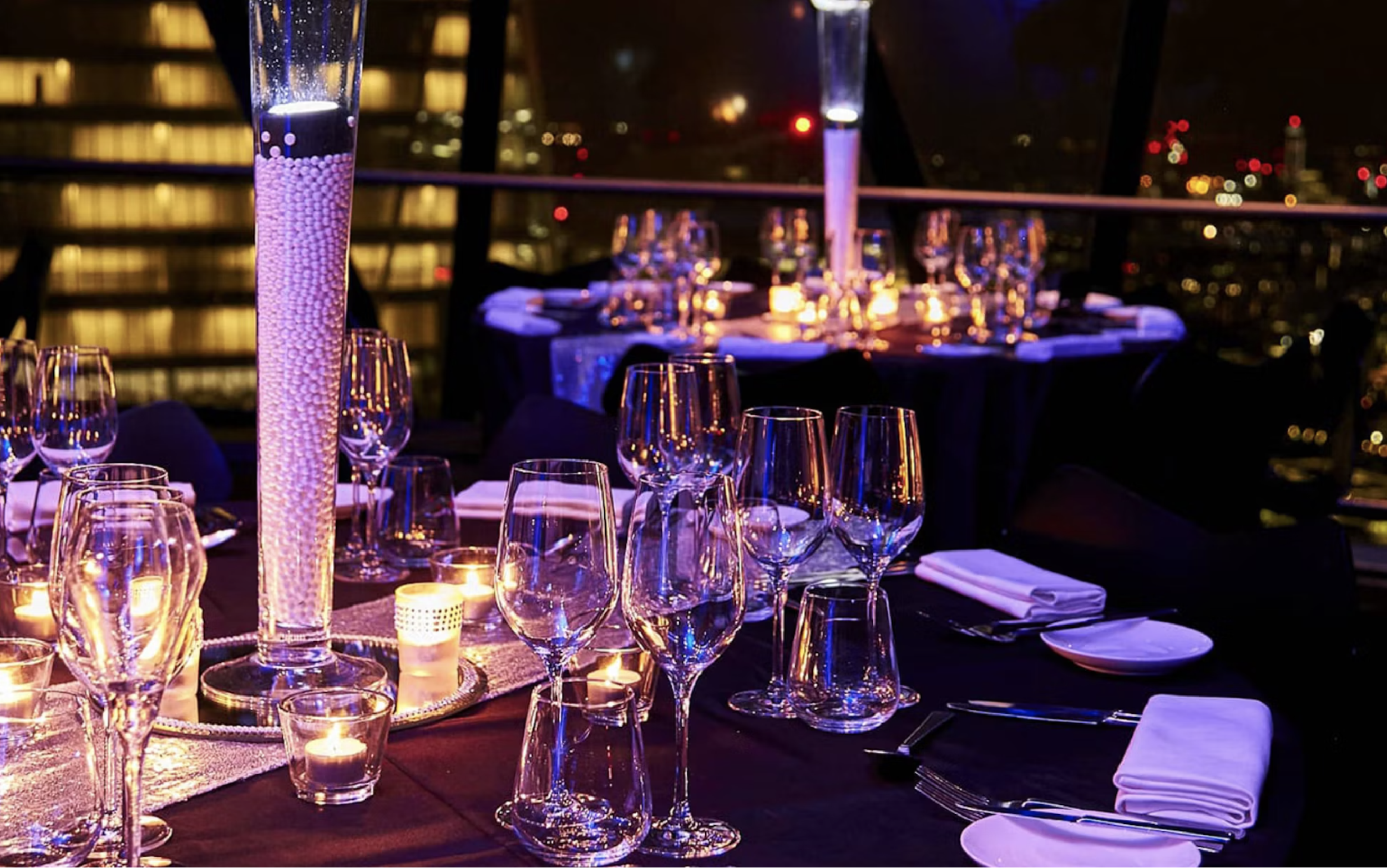 Elegant dining table set for an event with multiple wine glasses, candles, and a tall centerpiece, overlooking a city skyline at night.
